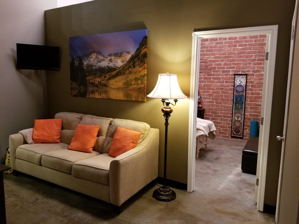 Small living room with beige sofa, orange pillows, wall art, and floor lamp leading to a brick-walled room.
