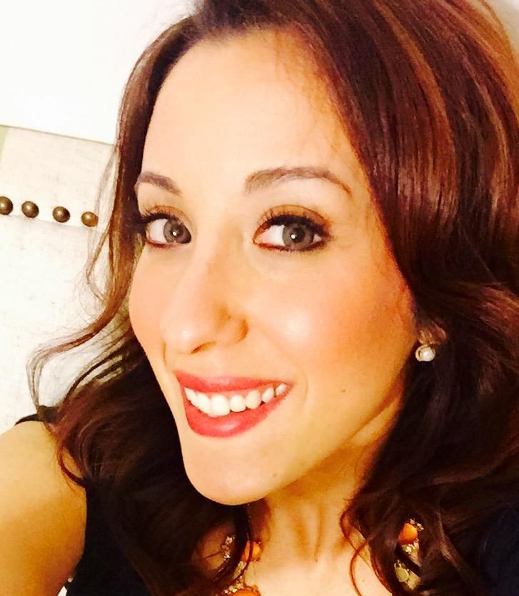 Close-up selfie of a smiling woman with wavy brown hair and red lipstick, against a white background