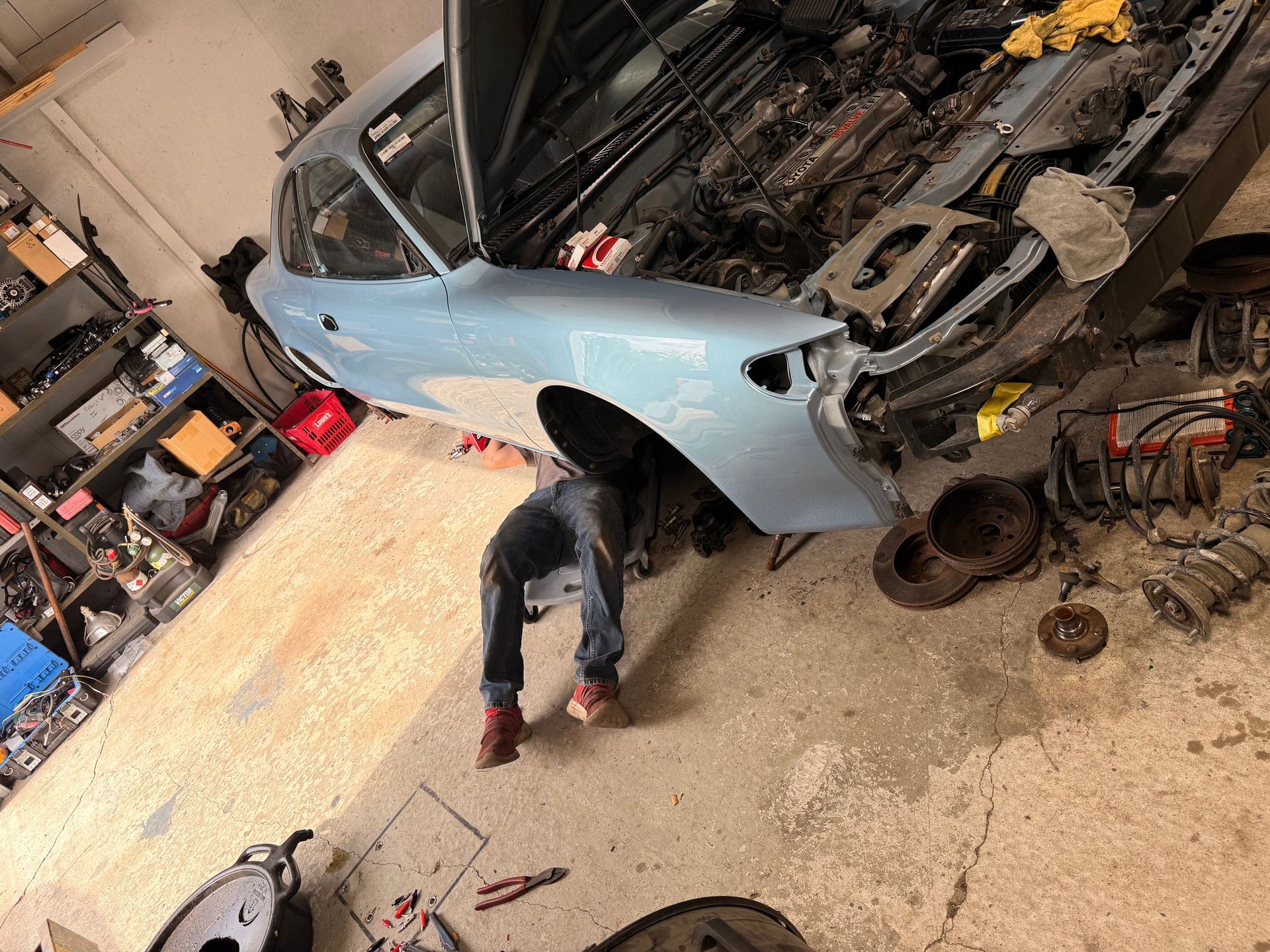 Person working on a light blue car in a cluttered auto repair garage