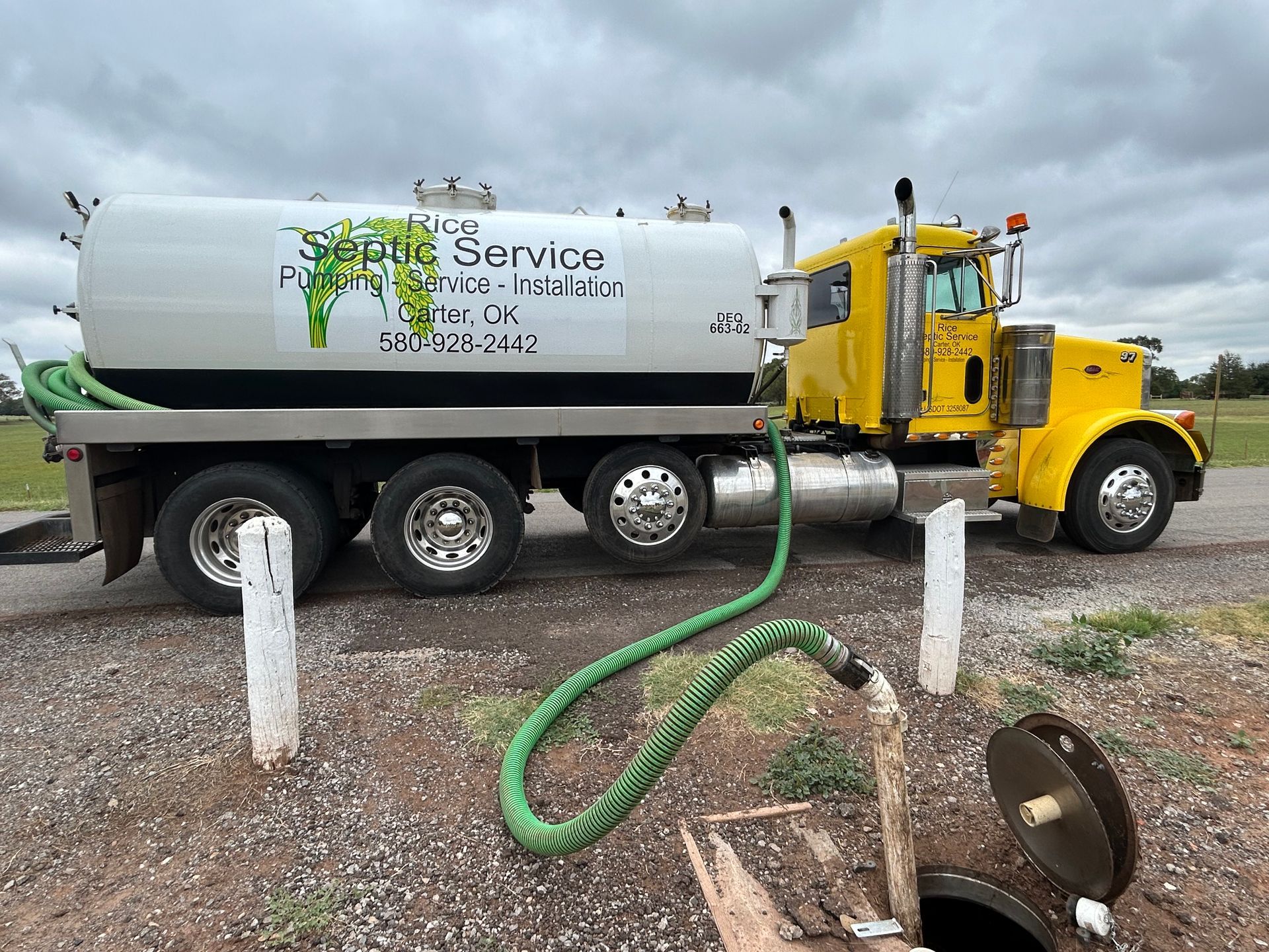 Yellow septic service truck with green hose pumping from a manhole on gravel. Cloudy sky.