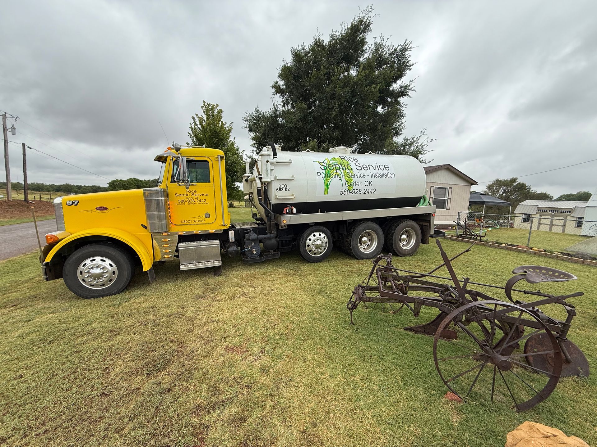 Yellow septic truck parked on a grassy field, near an old farm plow, under a cloudy sky.