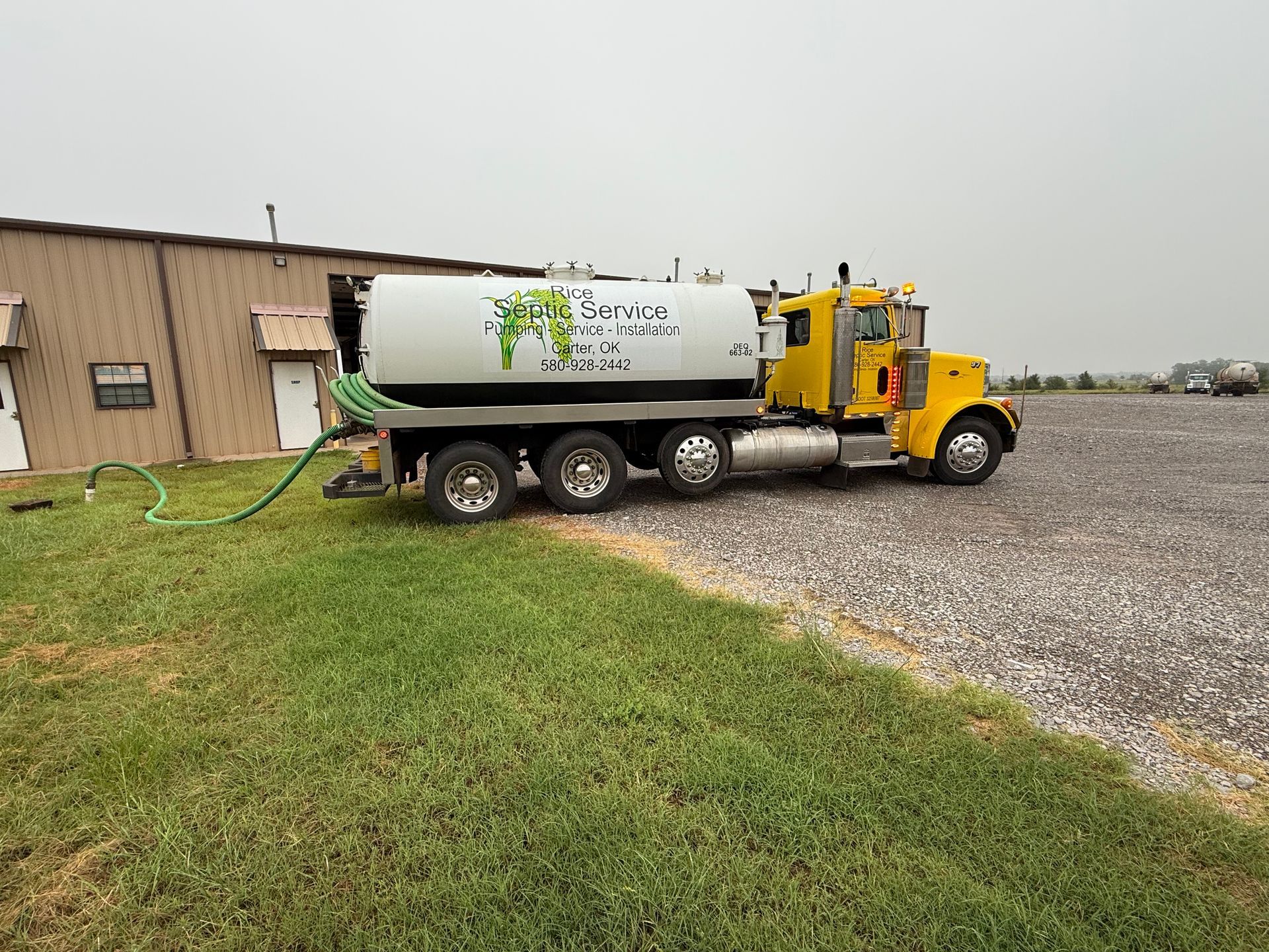 Yellow tanker truck parked by a building, connected to a hose on green grass, gravel ground.