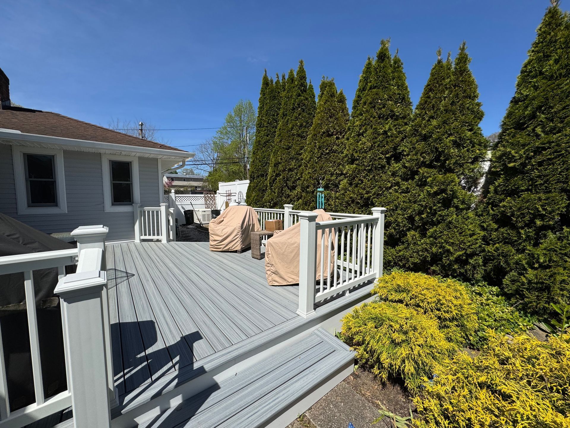 Gray deck with white railings, surrounded by greenery and a house on a sunny day.