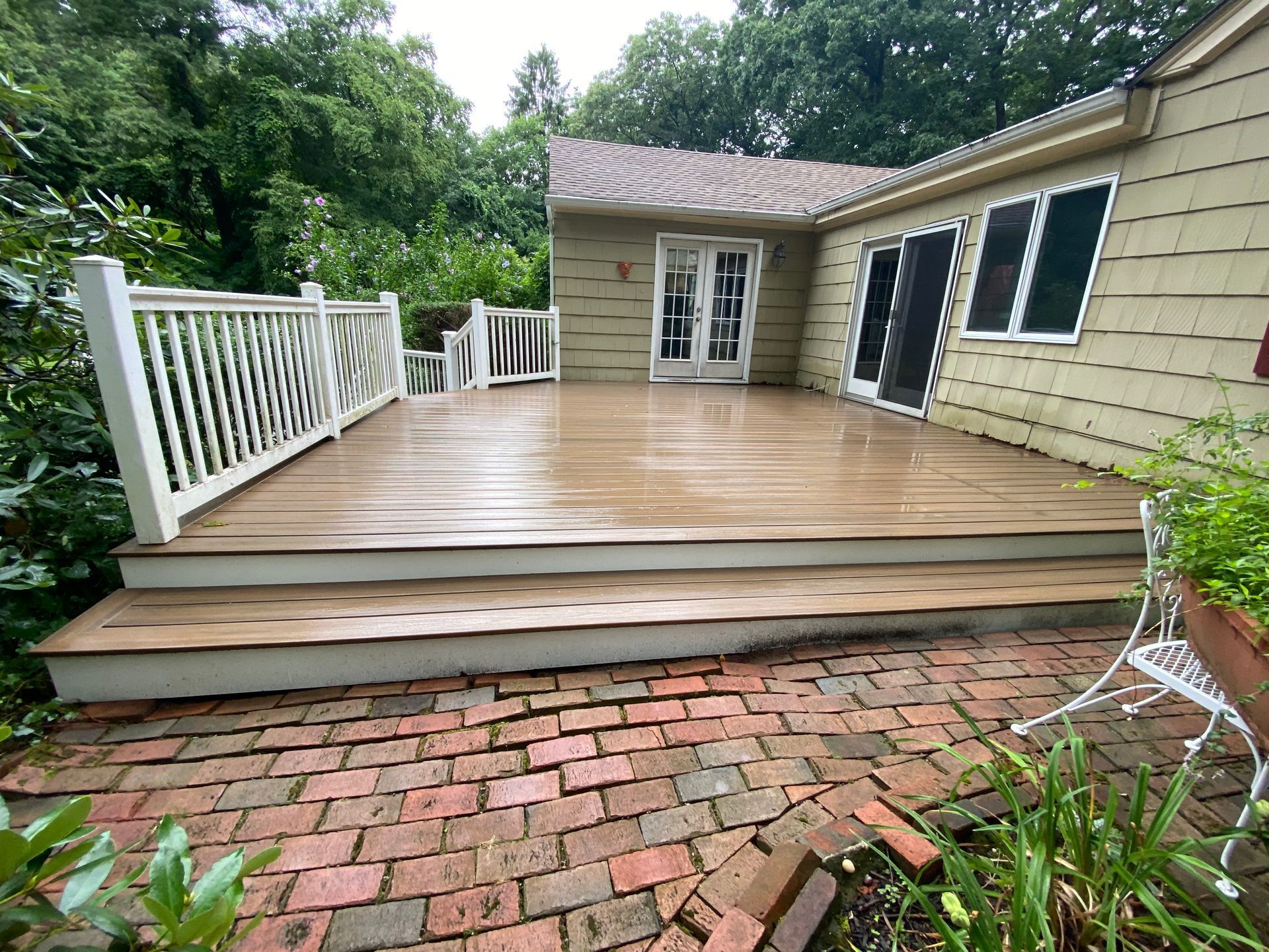 A wooden deck with white railing next to a house with a red brick patio.