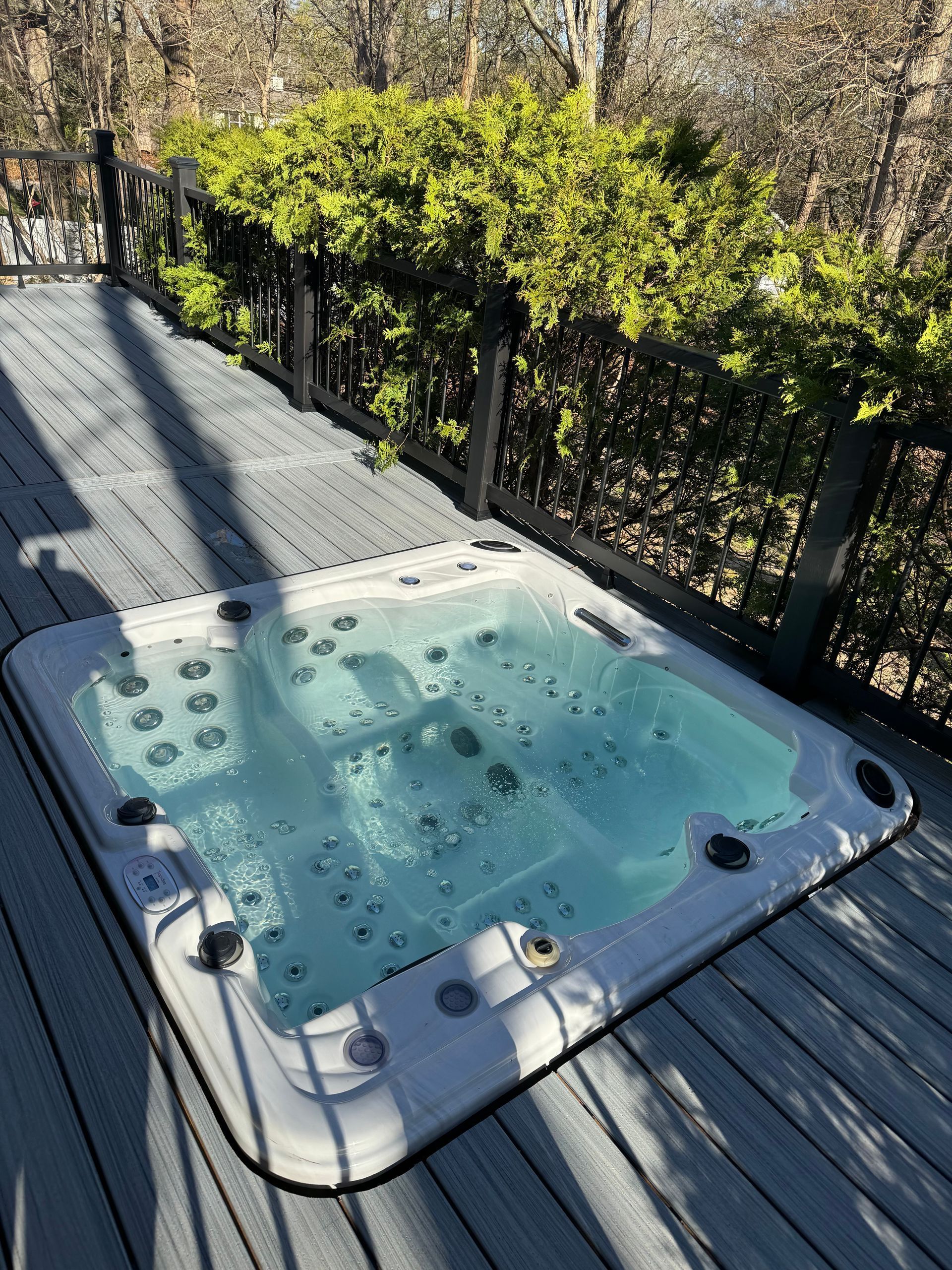 Hot tub on gray deck with black railing and green foliage in the background.