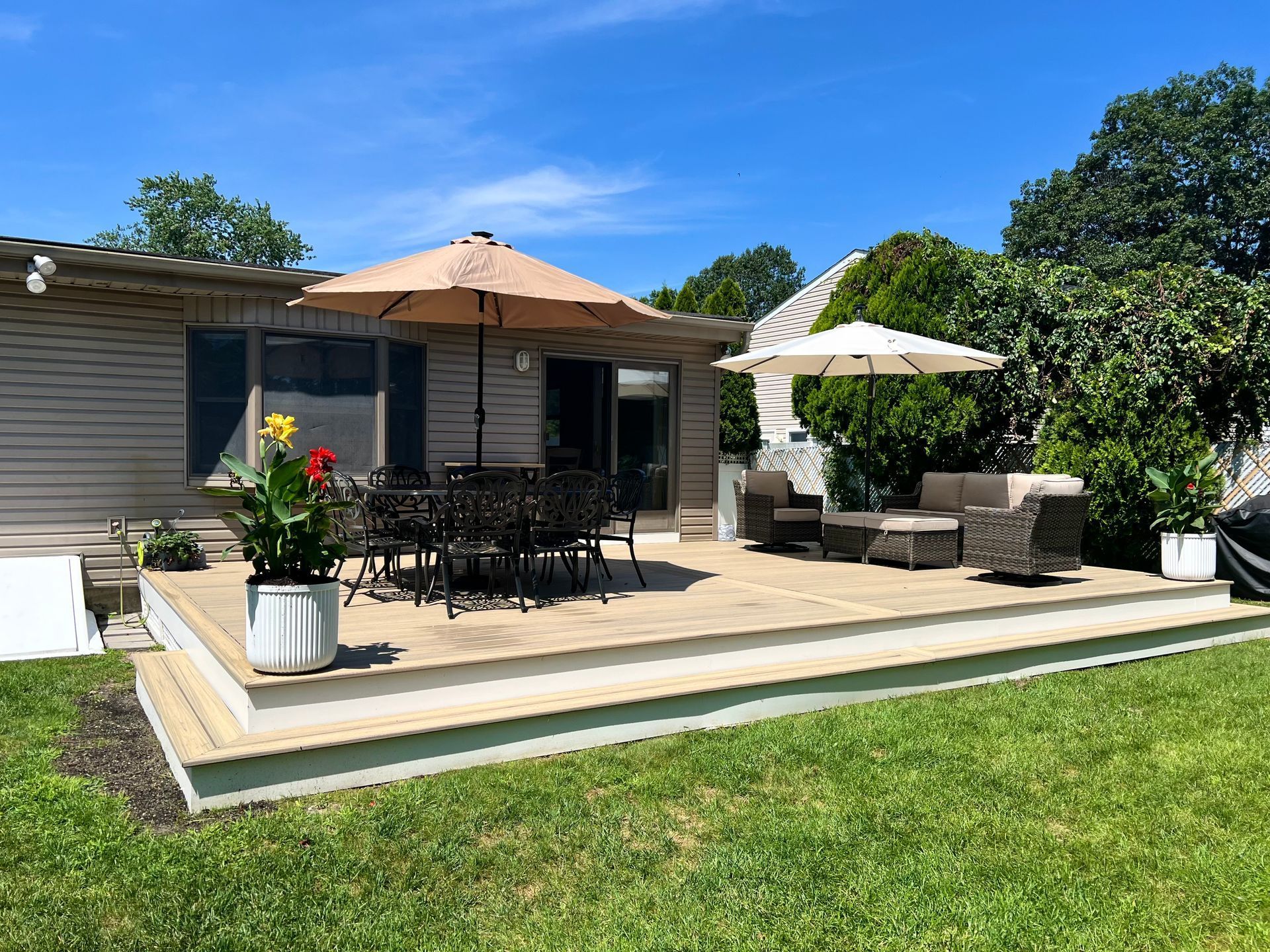 A deck with patio furniture and umbrellas, next to a house and grass on a sunny day.