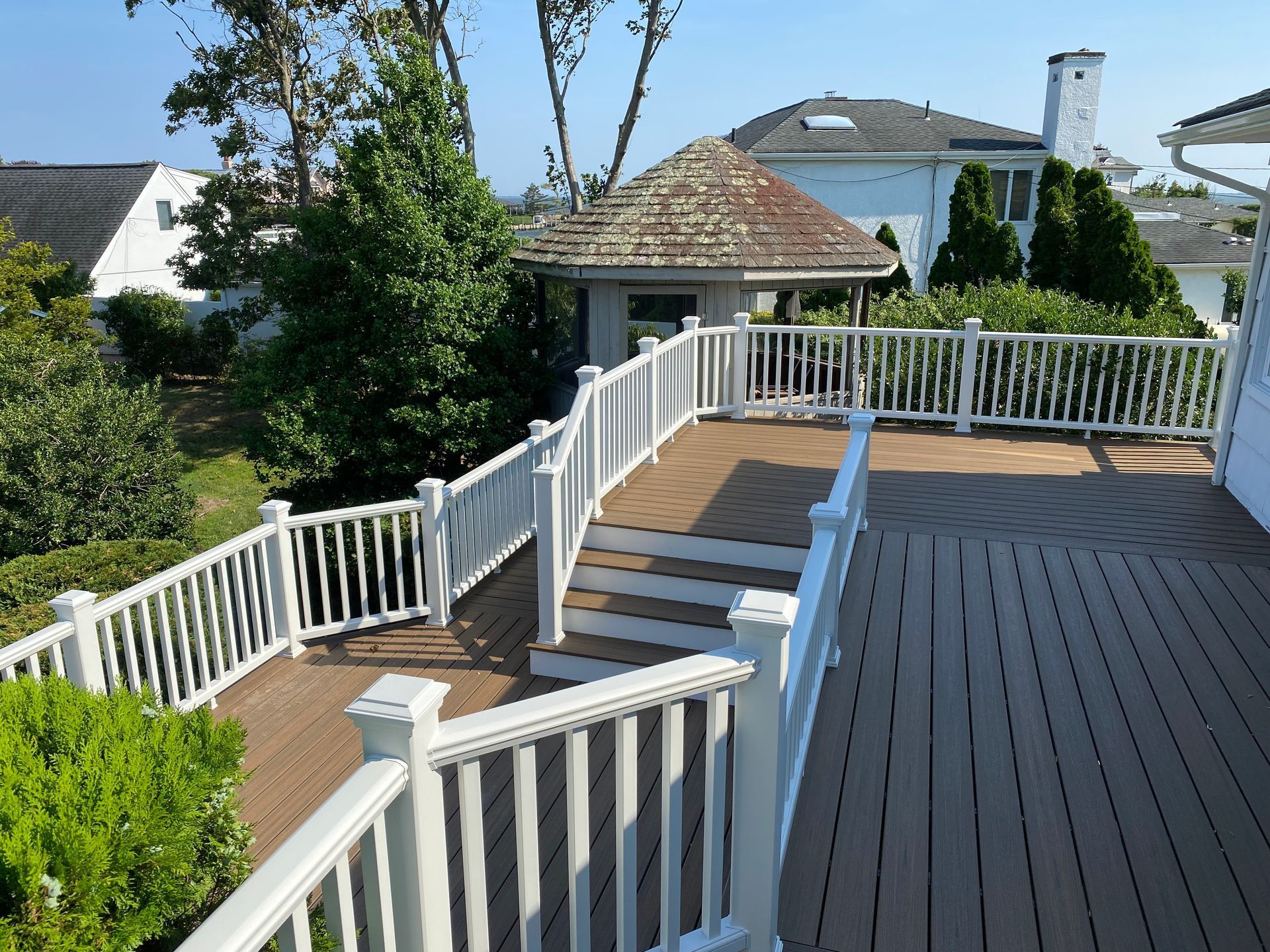 Wooden deck with white railings and stairs leading to a gazebo. Green trees and houses in the background.