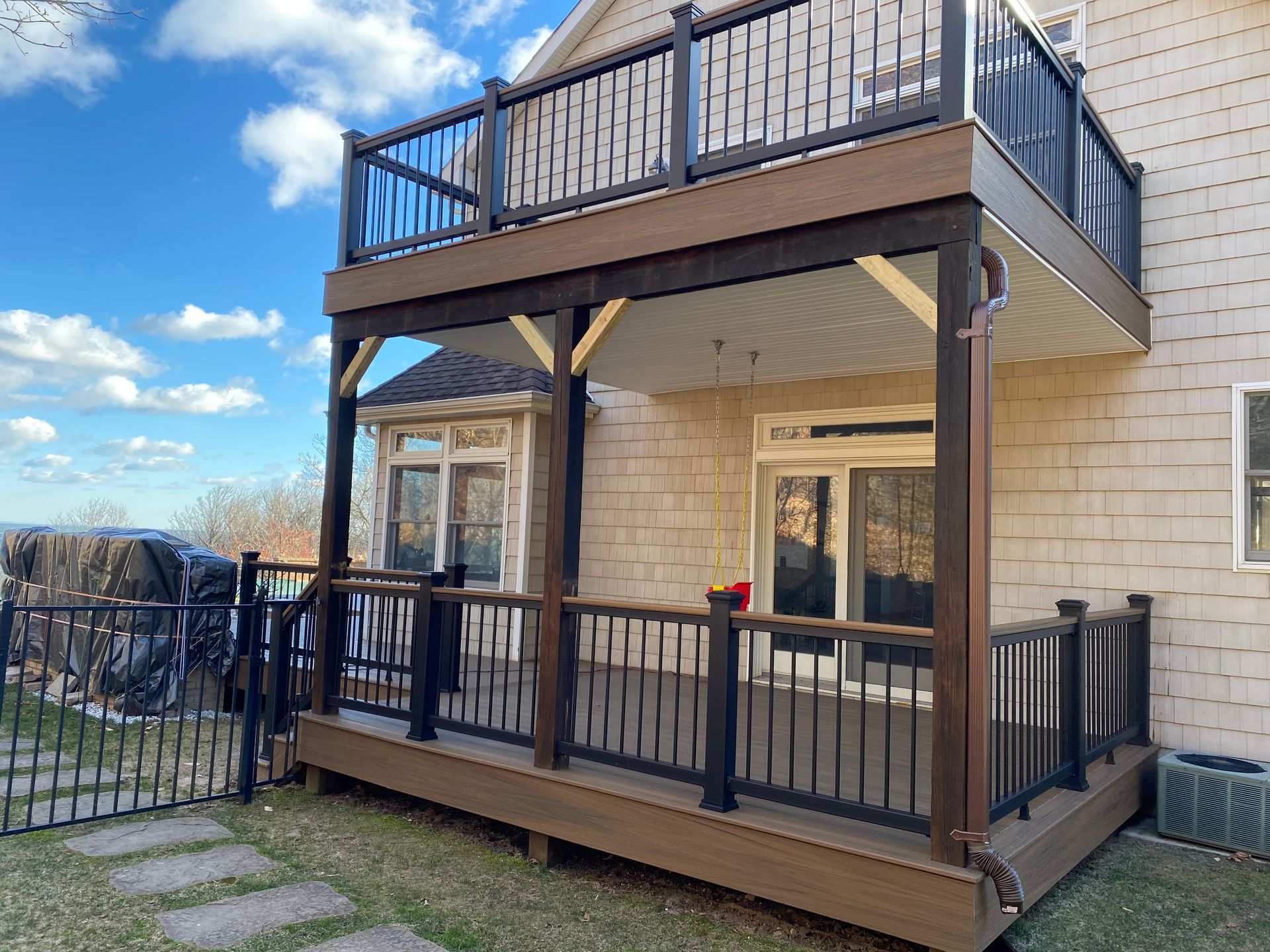 Two-story deck on a house, brown composite boards, black railings. Blue sky, white house.