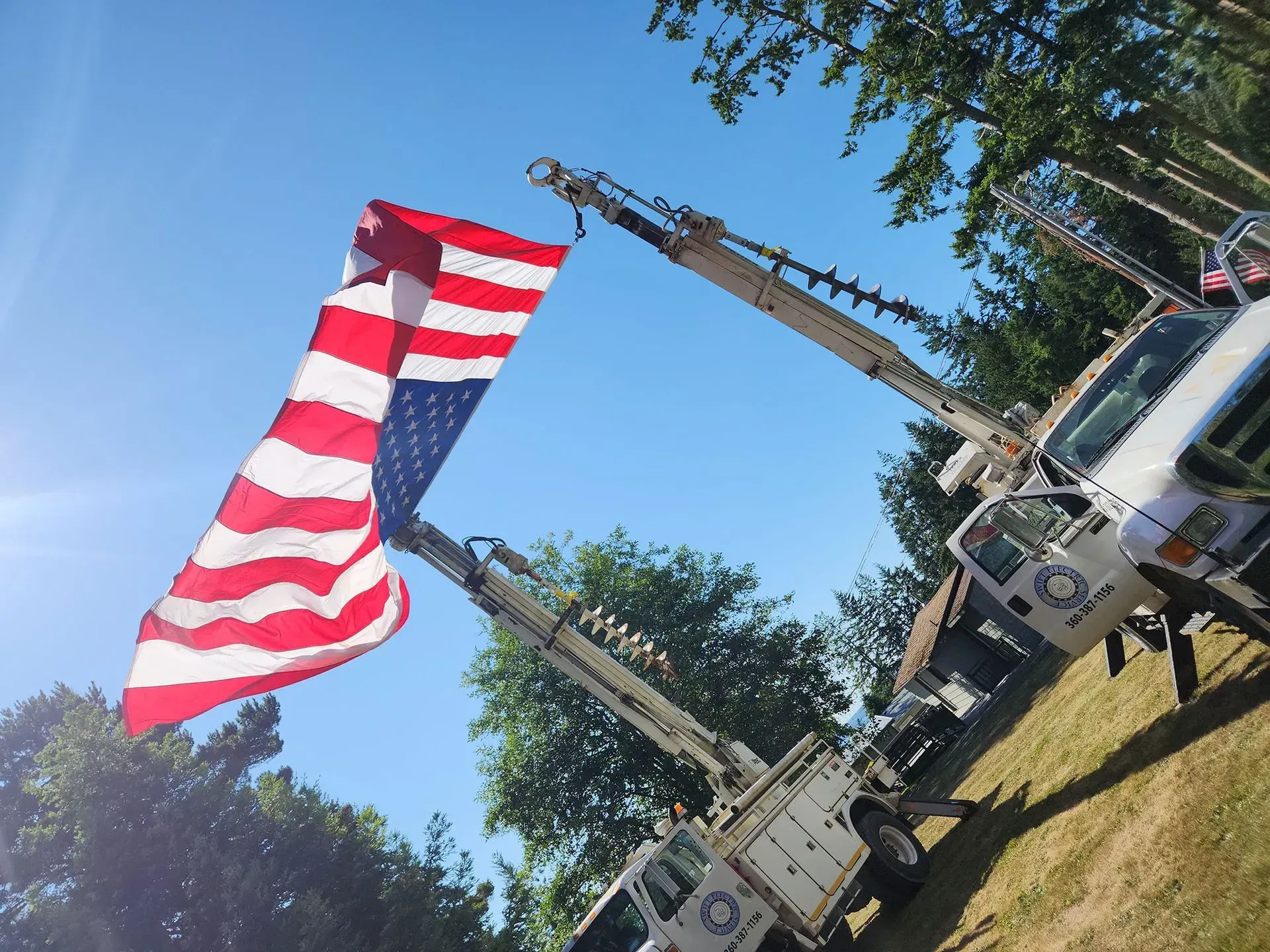 Two utility trucks with raised booms hold a large American flag against a blue sky.