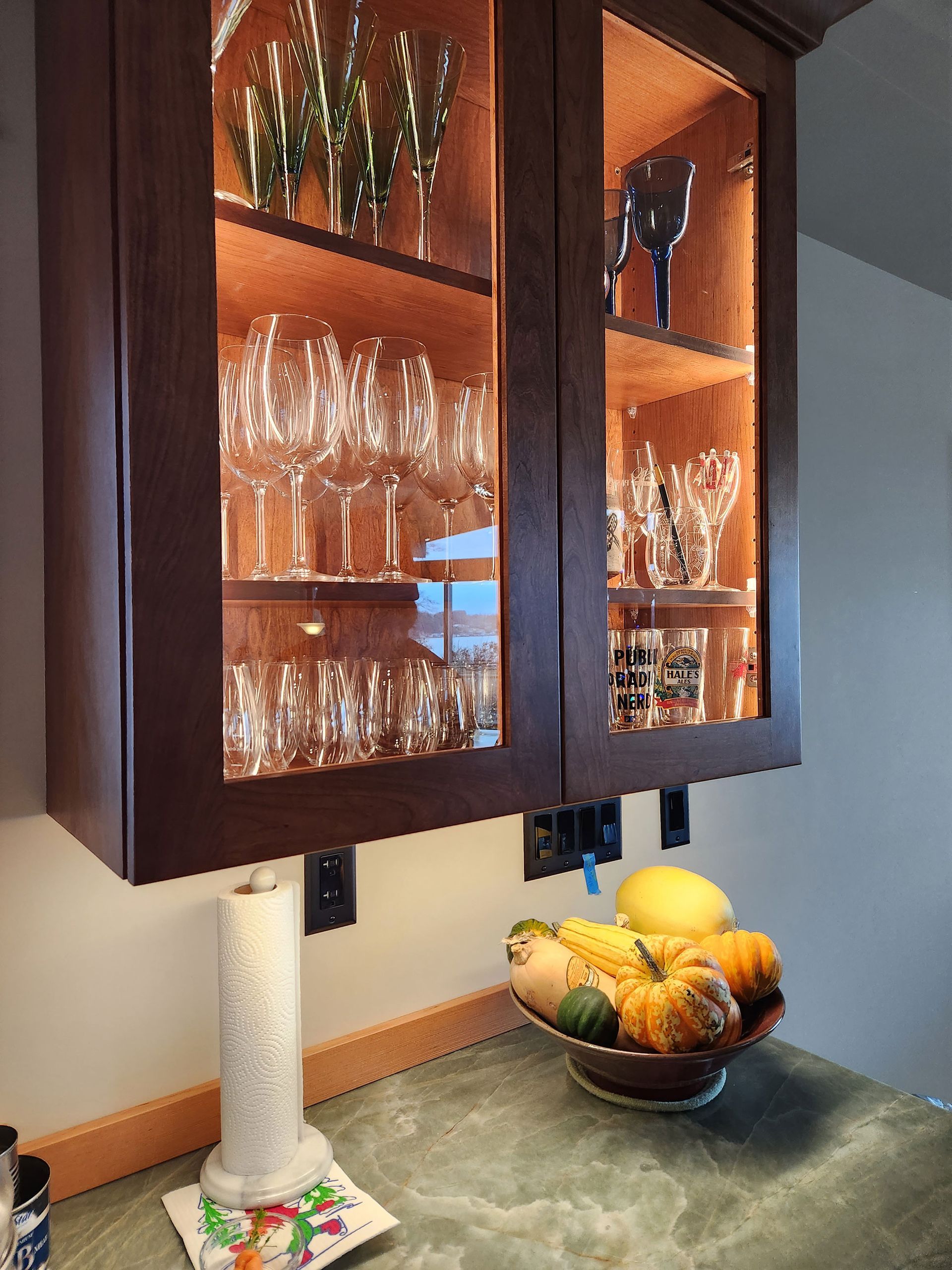 Dark-stained glass-front cabinets with interior lighting showcase glassware. A countertop holds a bowl of fruit and a paper towel holder.