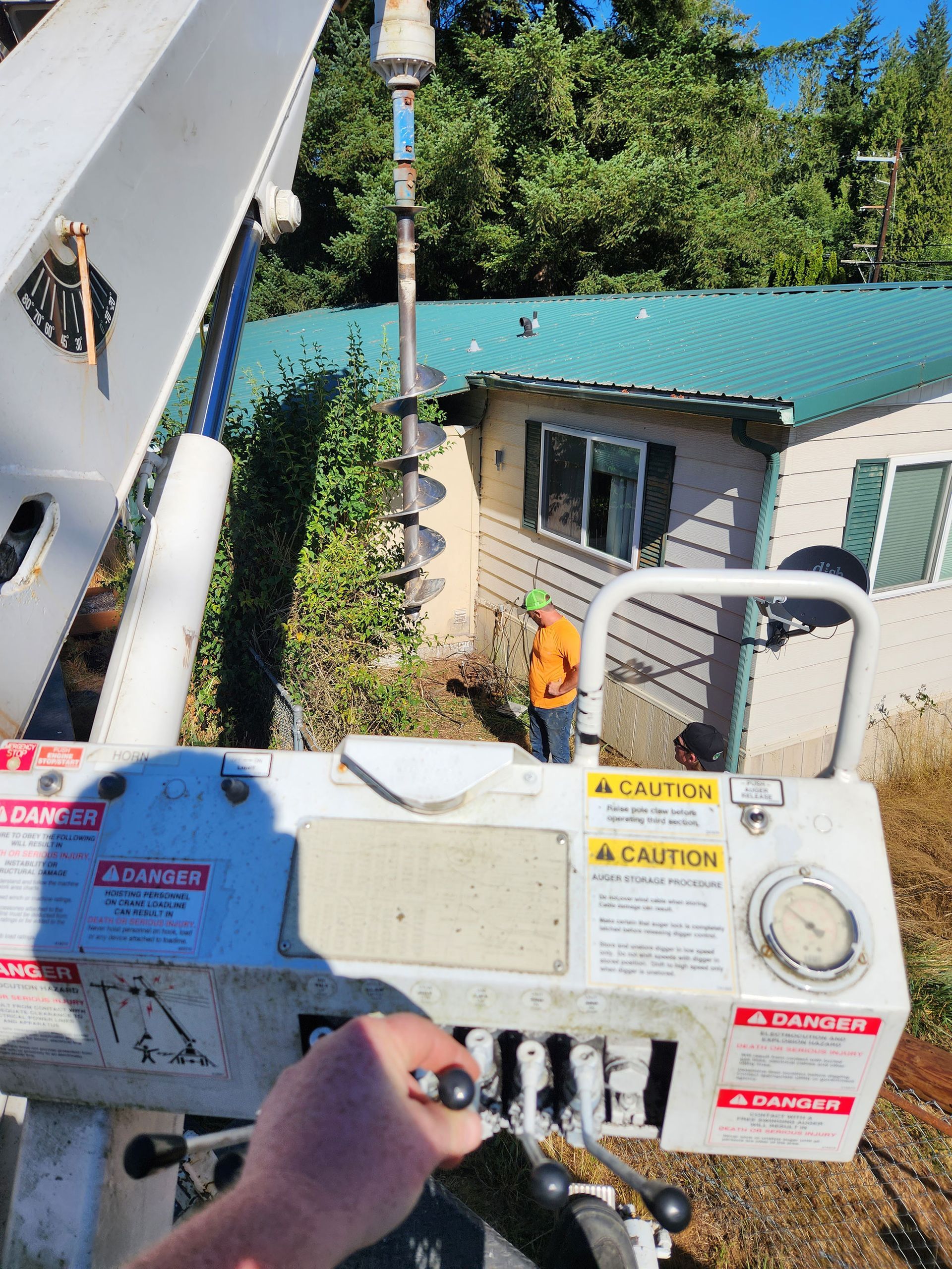 A person operating a drilling machine near a house with green roofing; another person watches.