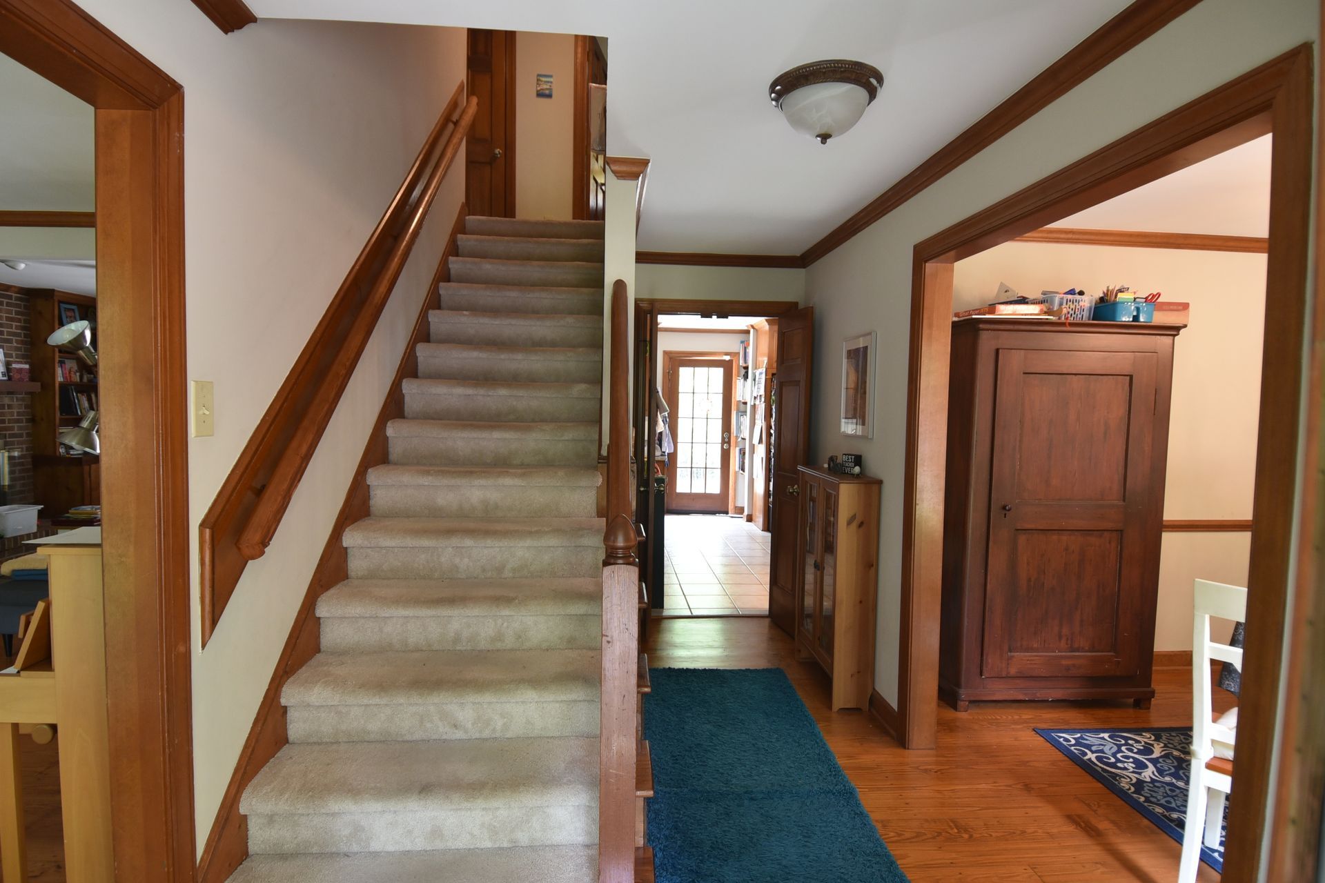 Staircase with light carpet and wooden banister. Hallway with hardwood floor, leading to other rooms.