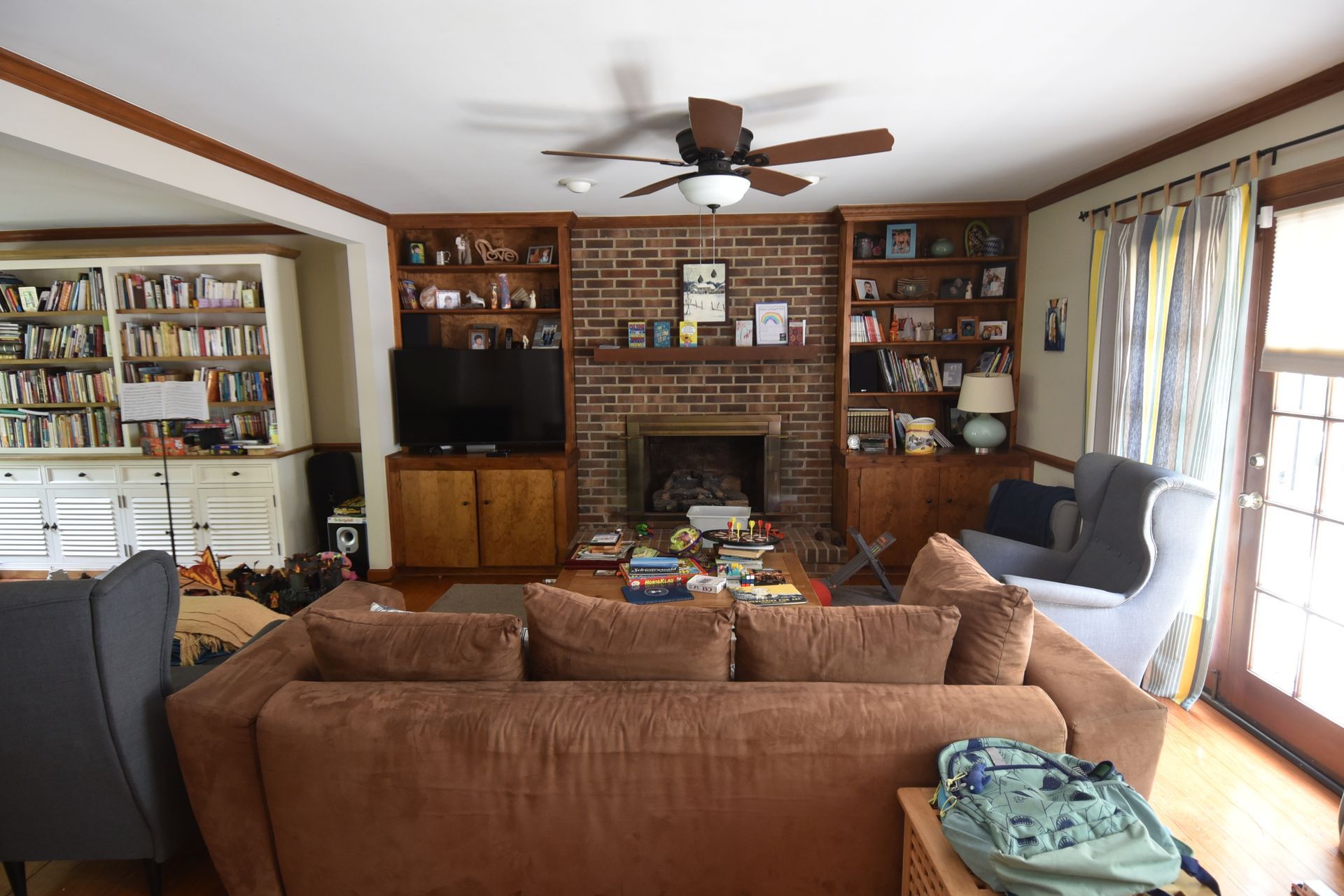 Living room with brown couch, brick fireplace, built-in bookshelves, and a ceiling fan.