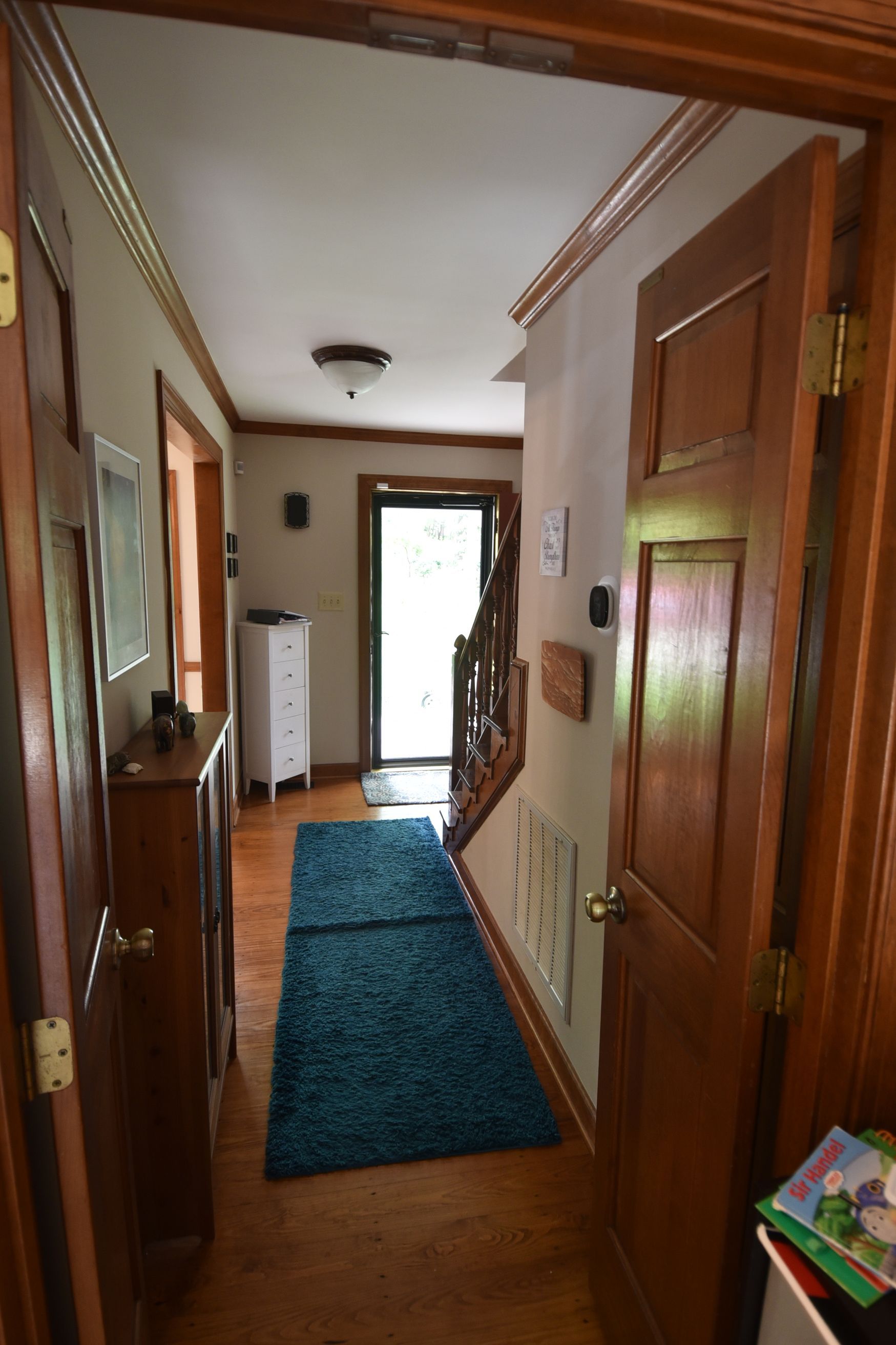 Narrow hallway with wood floor, blue rug, wood doors, leading to a lit doorway and stairs.