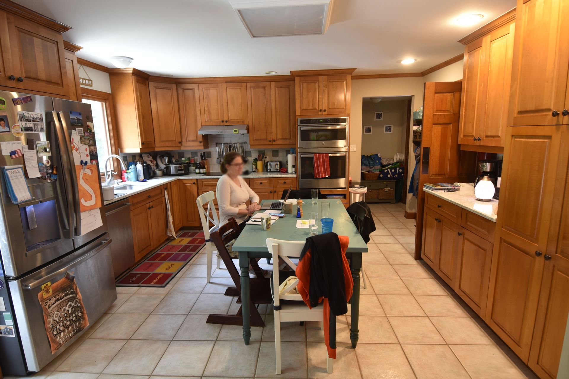 Kitchen with wooden cabinets, a person at a table, stainless steel fridge, and tile floor.