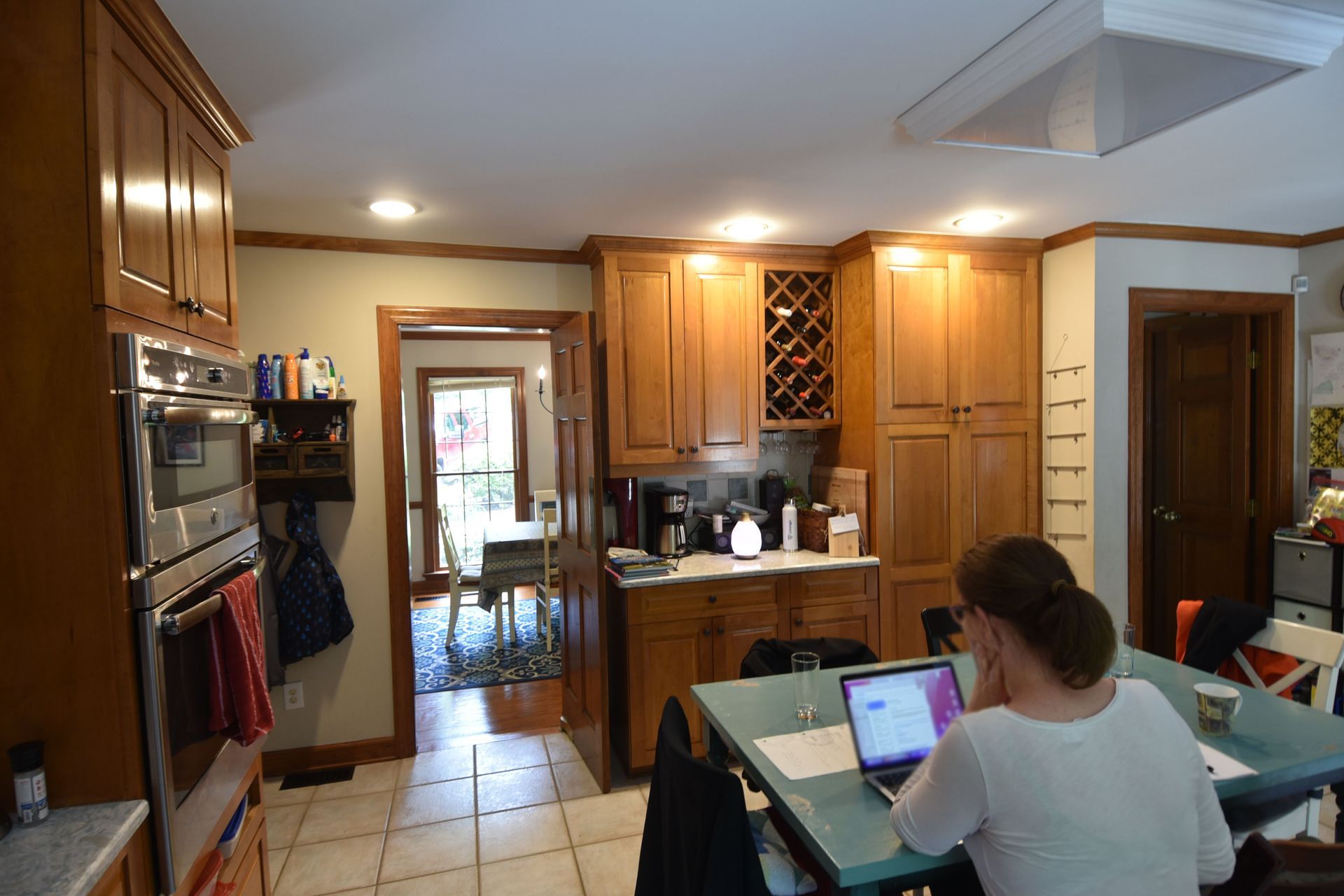 A person works on a laptop at a kitchen table. Wooden cabinets, oven, and a doorway are visible.
