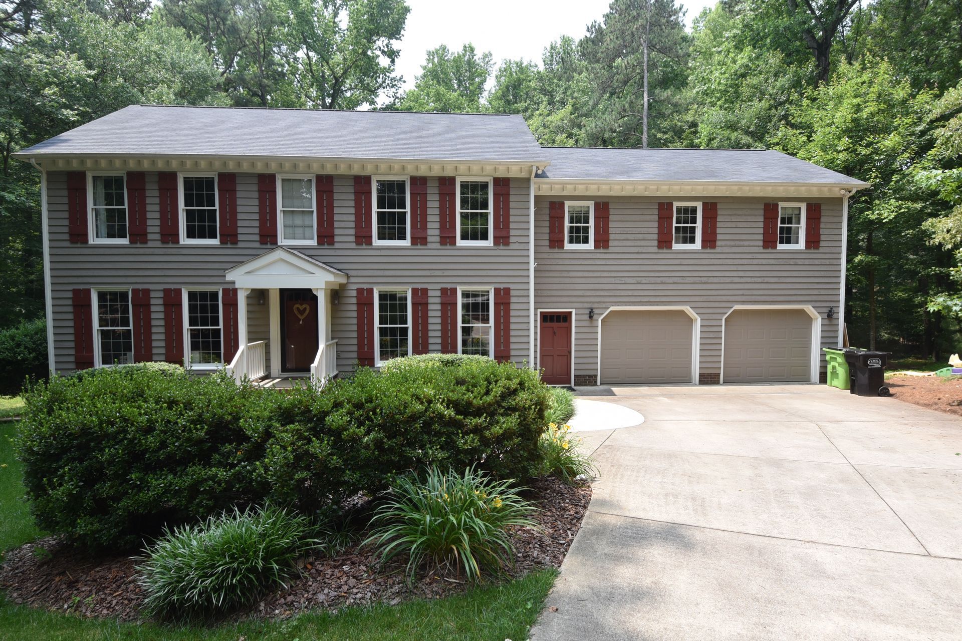 Two-story gray house with burgundy shutters, two-car garage, and driveway. Green bushes in front.