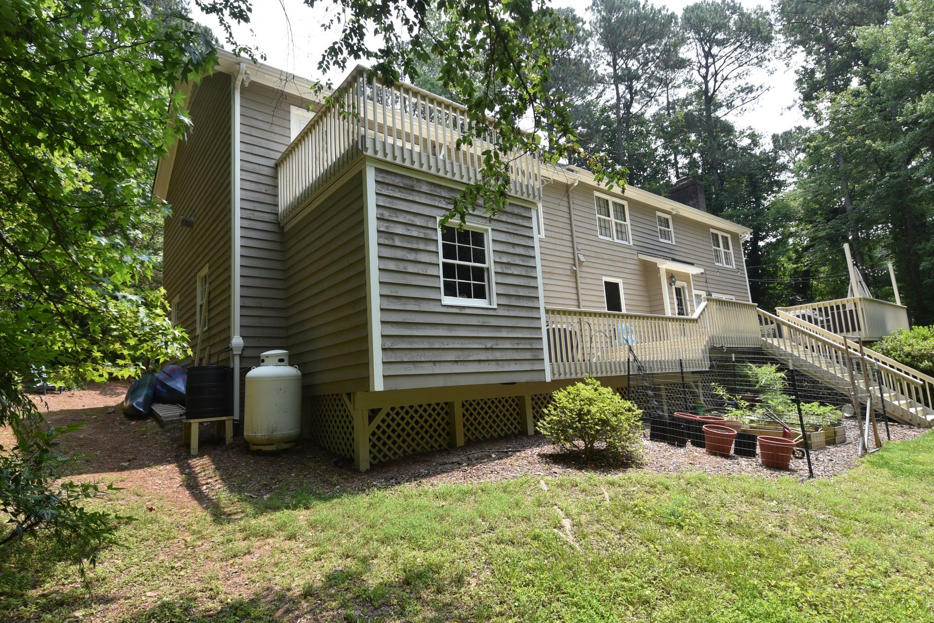 Beige house with wooden deck and stairs, surrounded by grass and trees.