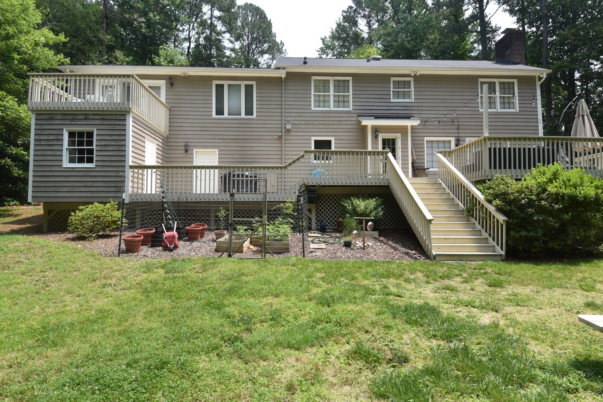 Back of a two-story house with wooden decks and stairs, tan siding, green grass, and trees in the background.