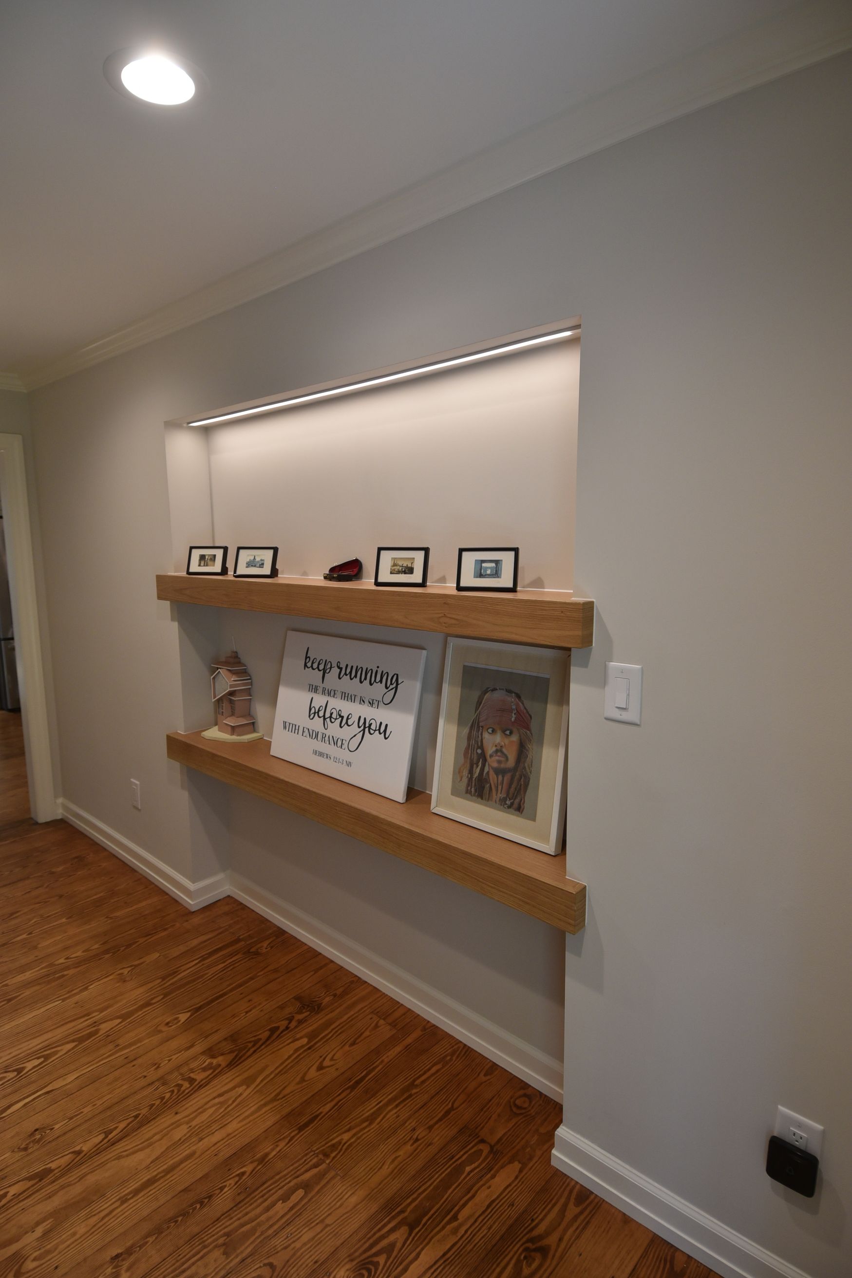 Built-in wooden shelves in a recessed wall niche, displaying framed photos and decor, lit by a recessed light.