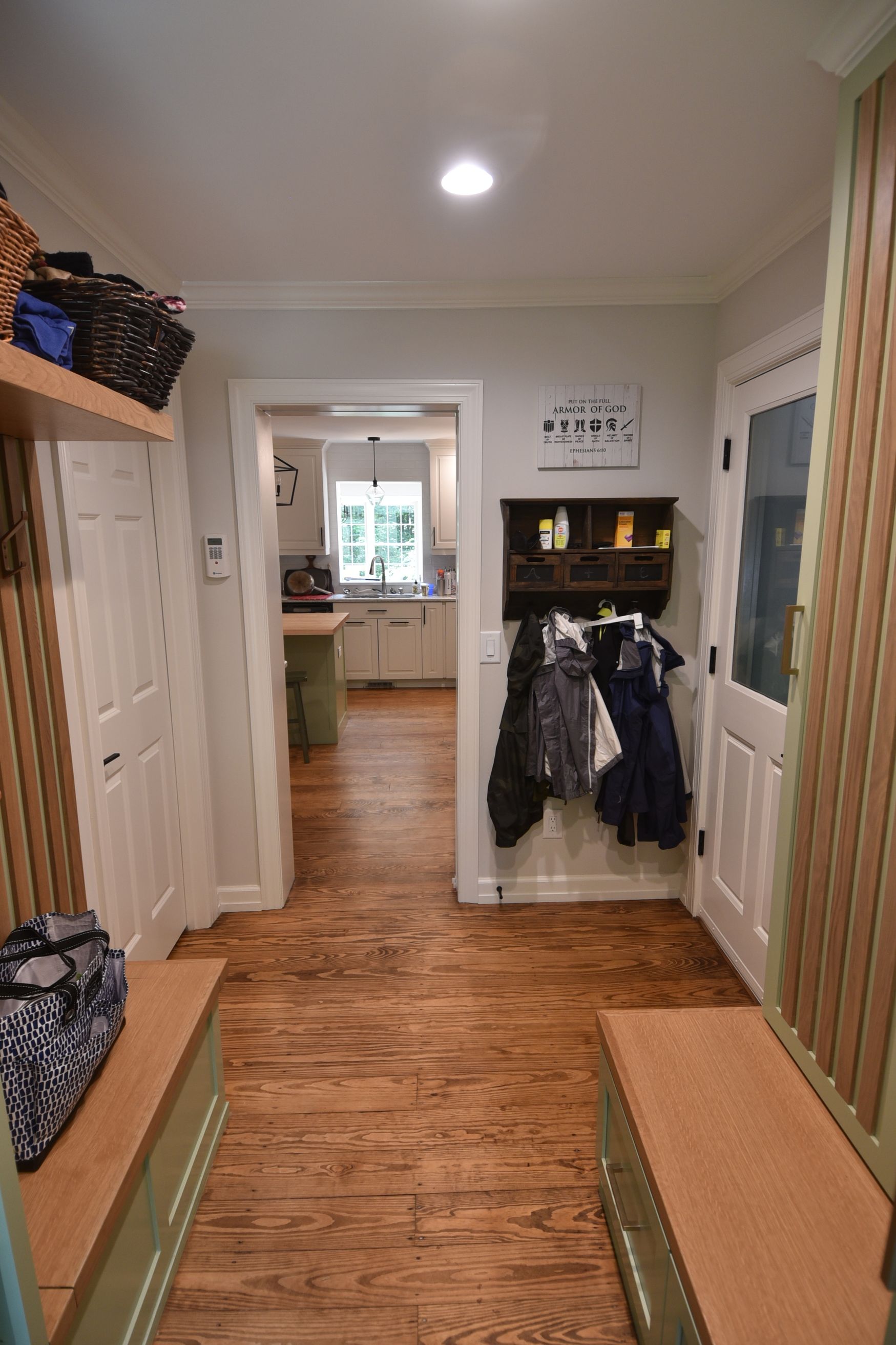 Entryway with wooden floor and benches, coats hanging on a wall, and a view into a kitchen.
