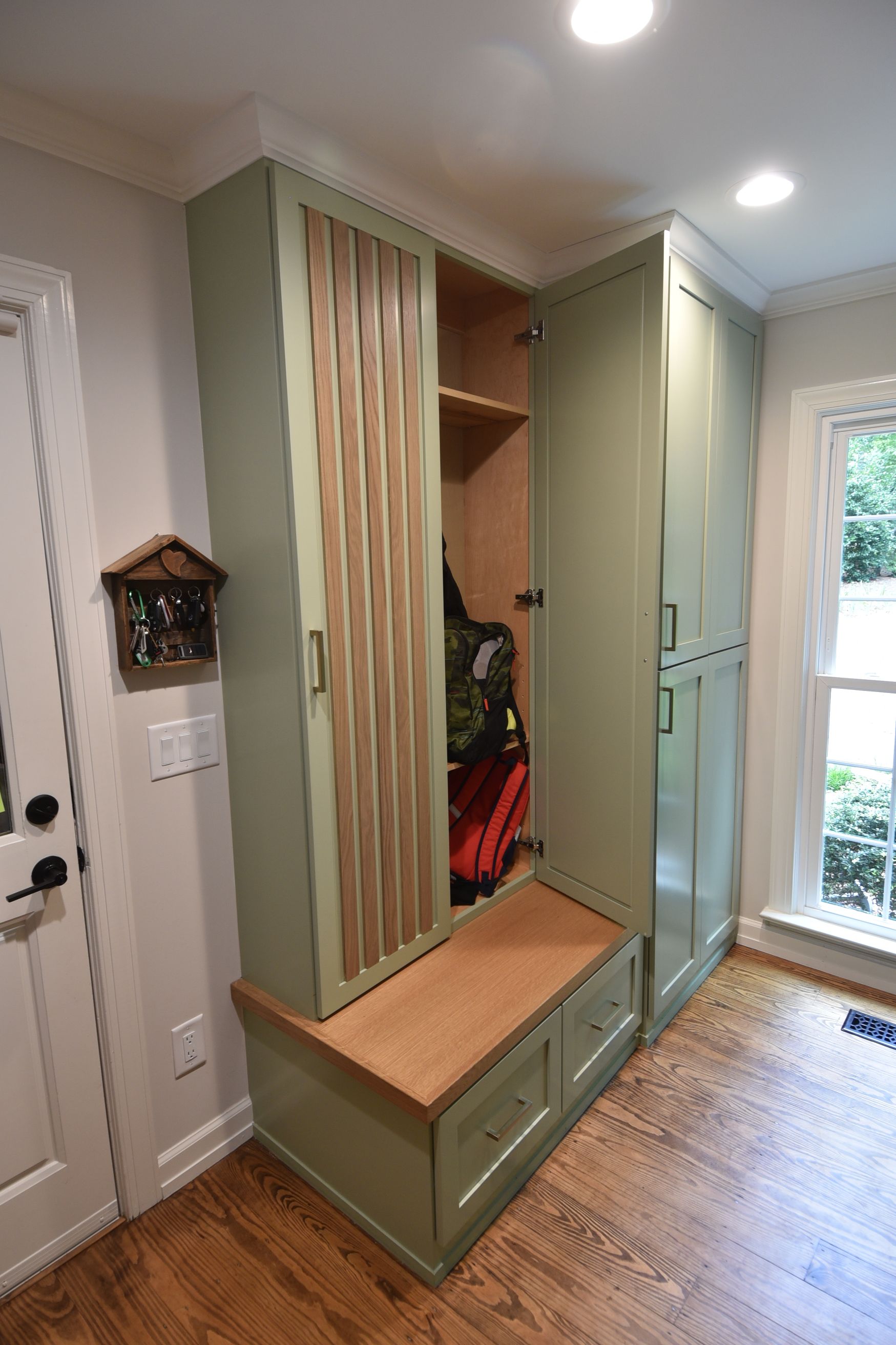 Green built-in storage unit with bench, drawers, and tall cabinets. Door is open to reveal coats.