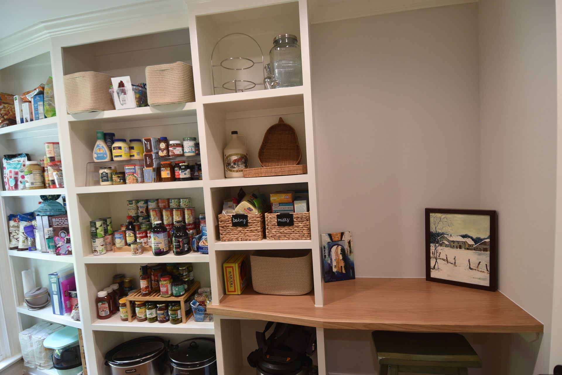 Built-in pantry with shelves stocked with food, and a countertop with a picture, over two garbage cans.