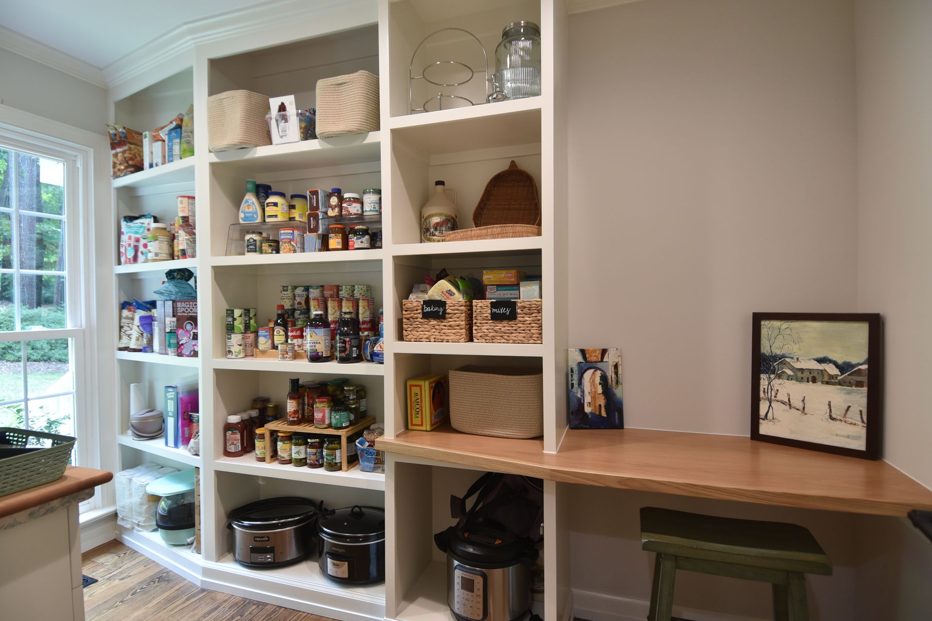Built-in pantry with white shelves, stocked with food. A wooden desk has a stool.