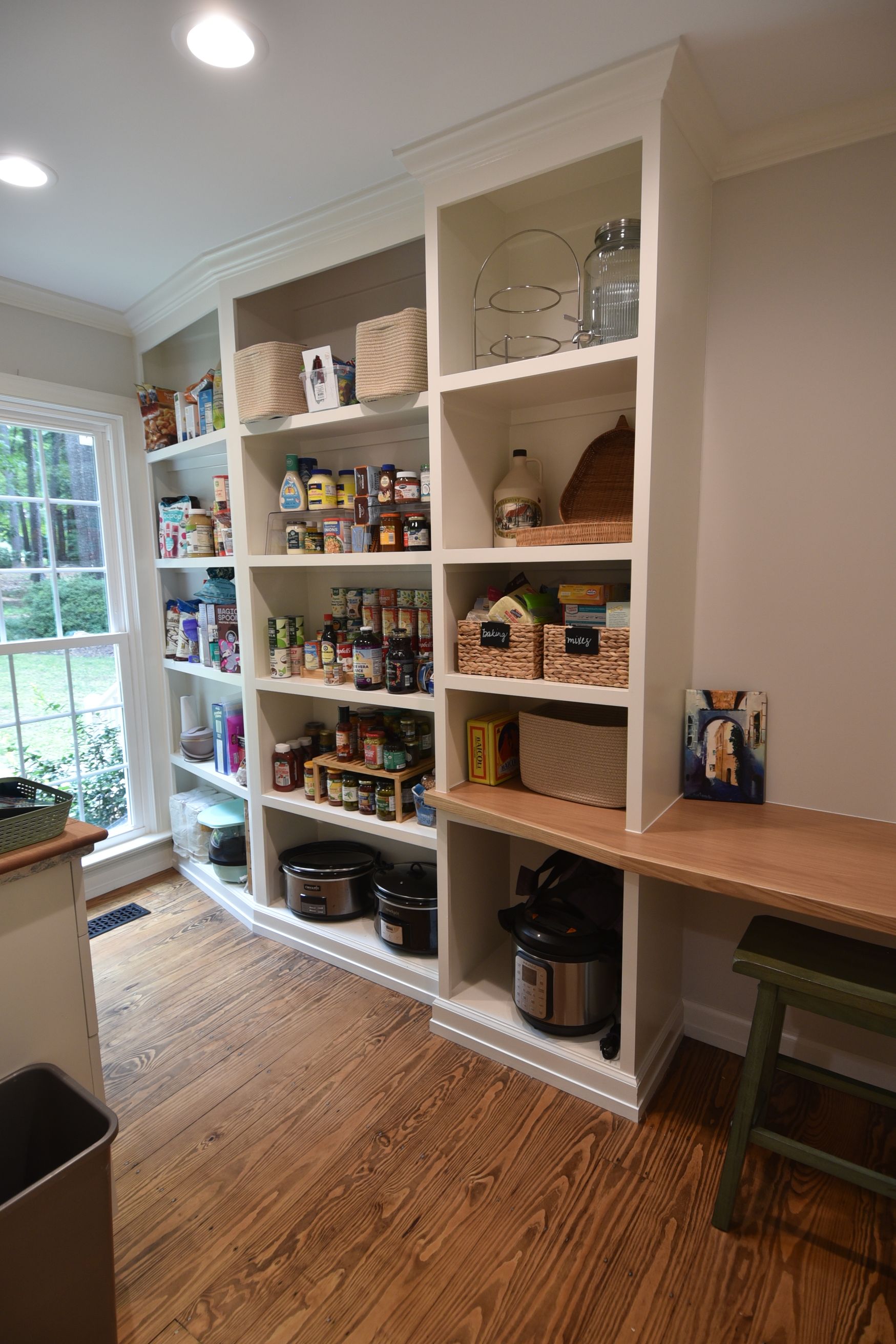 Pantry with white shelves filled with food, a built-in desk, and hardwood floors.