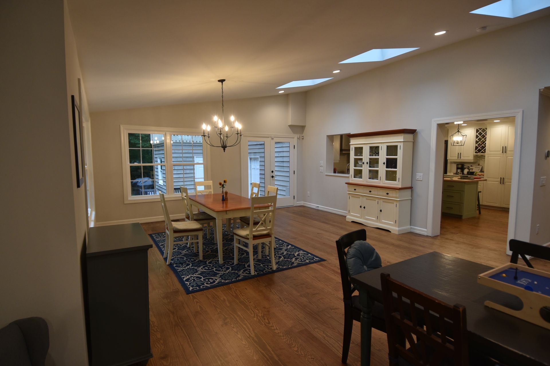 Spacious dining area with wood floors, table, chairs, chandelier, and light-filled kitchen in the background.