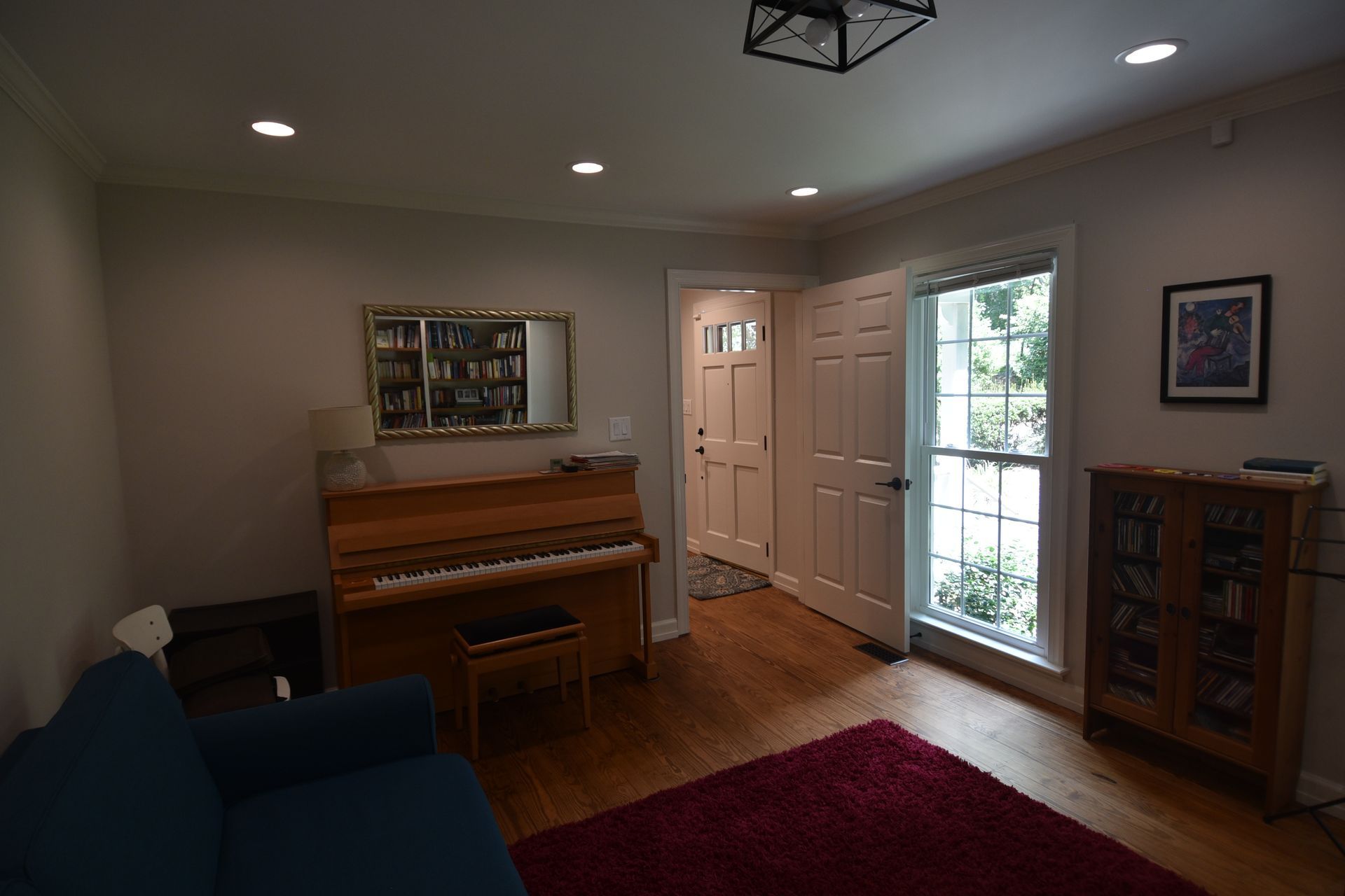 A room with a piano, blue couch, and burgundy rug. A door and window are visible.