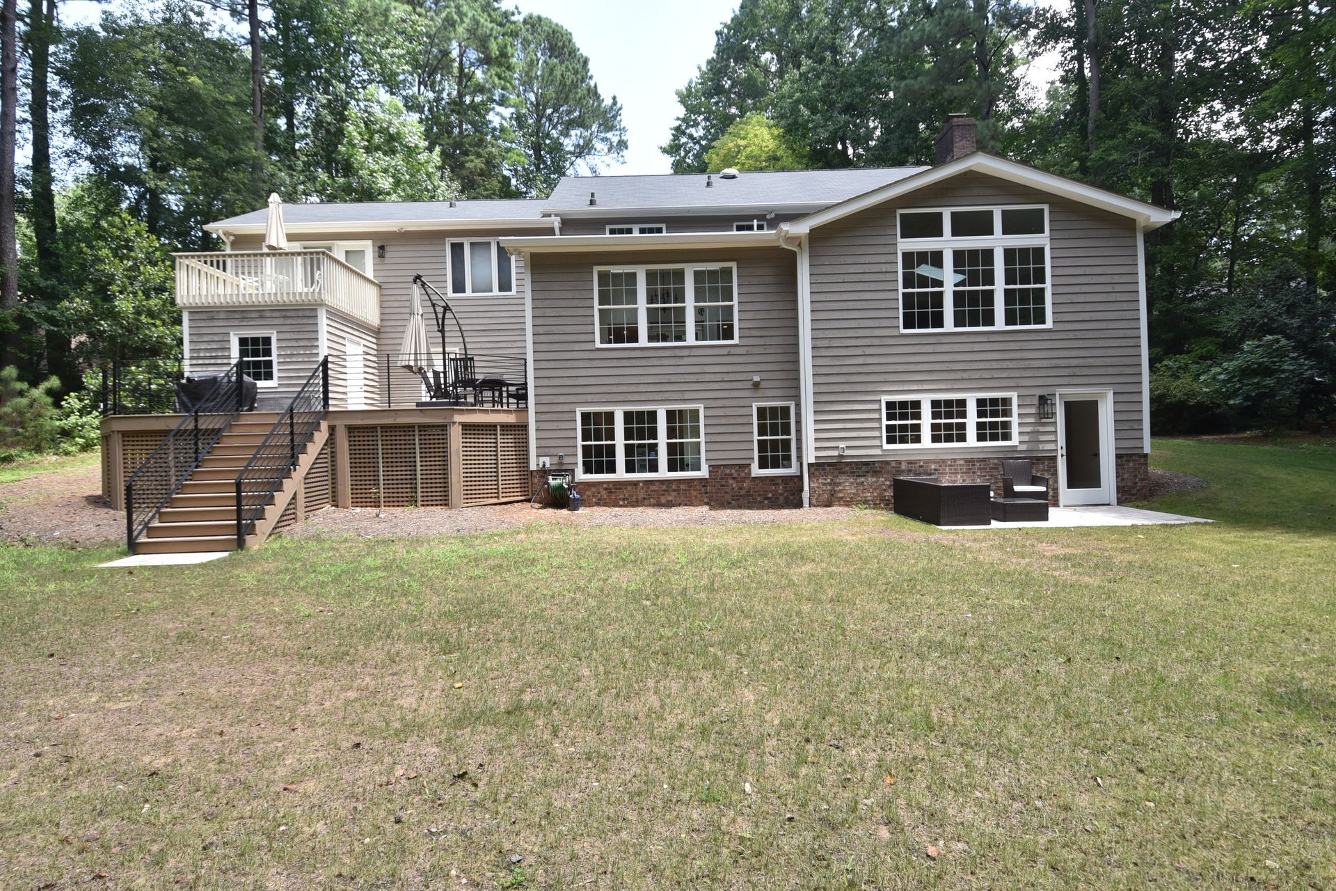 Back view of a two-story house with a deck, brown siding, and grassy yard in a wooded setting.
