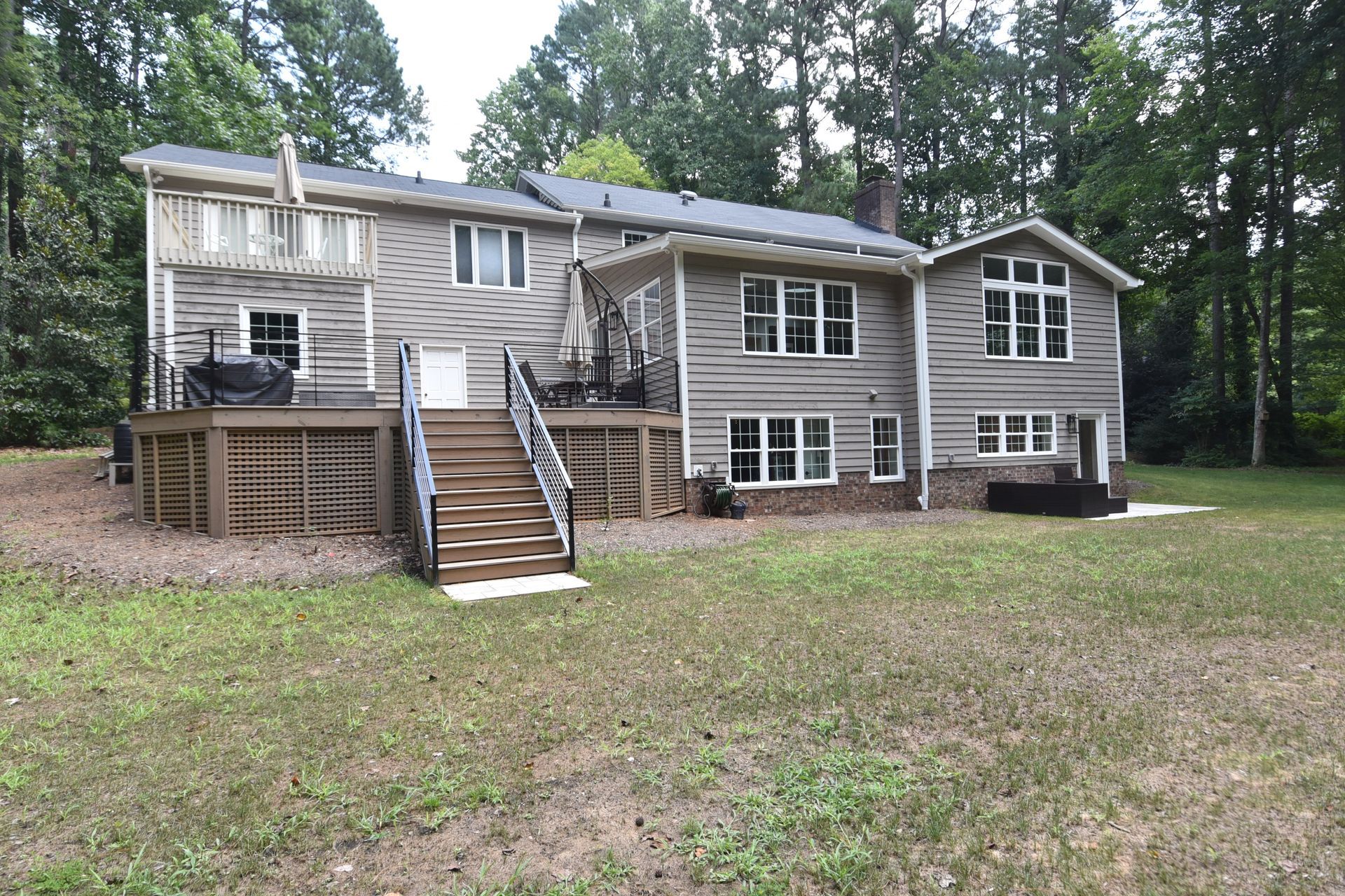 Back of a two-story gray house with a deck and stairs in a wooded backyard.