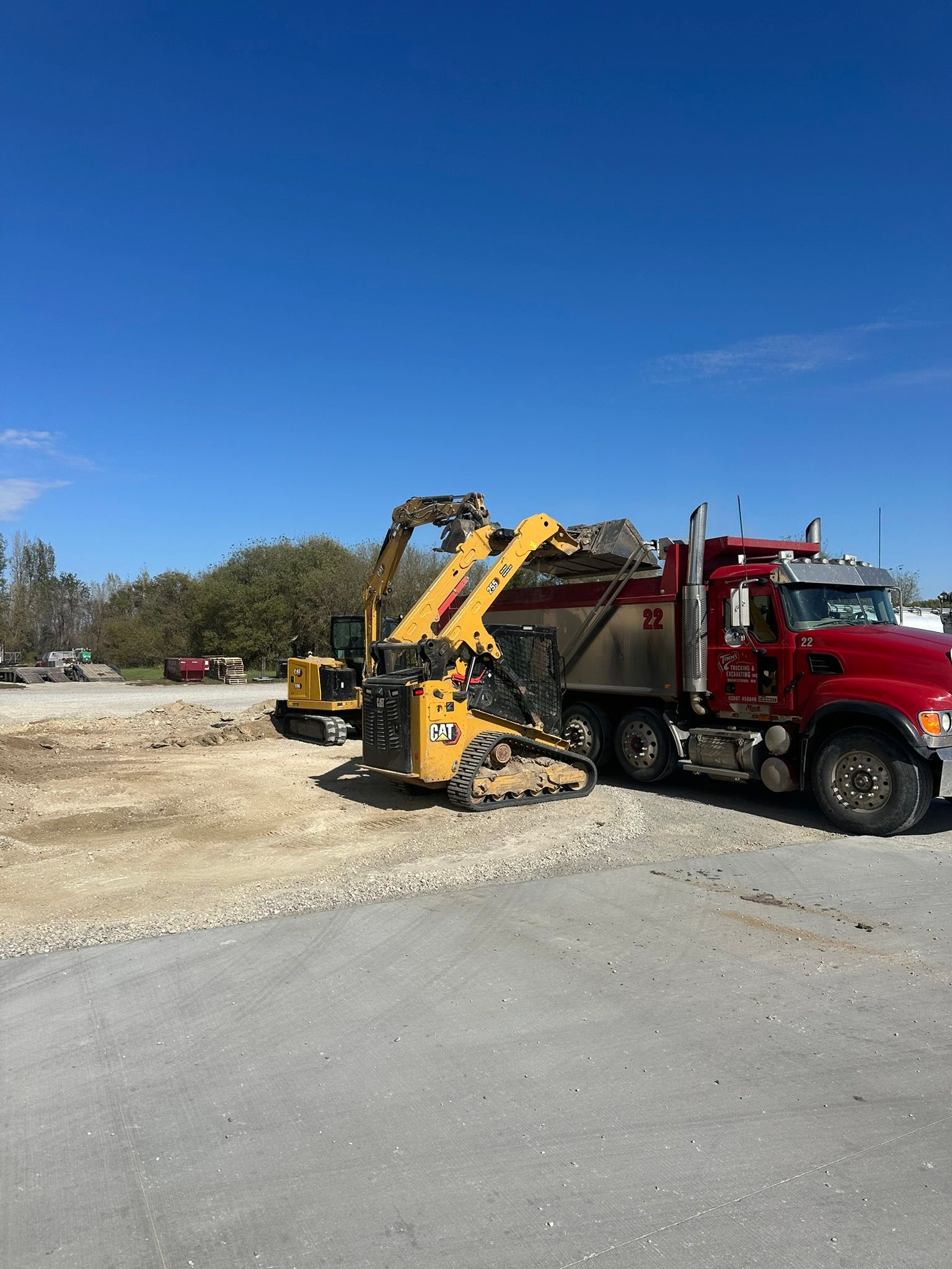 Yellow skid steer loading gravel into a red dump truck, both on a construction site. Blue sky.