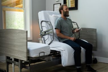A person sits on a hospital bed, holding a remote control, with the head of the bed partially elevated in a room.