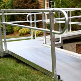 An outdoor aluminum wheelchair ramp with handrails, leading toward a house entrance with greenery in the background.