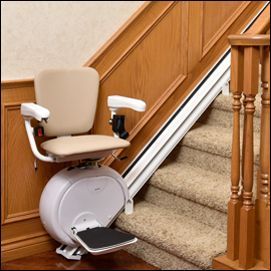 A beige stairlift mounted on a track alongside a staircase with light wood paneling and a wooden banister.