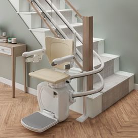 A cream-colored stairlift mounted on a curved rail at the base of a white wooden staircase in a home interior.