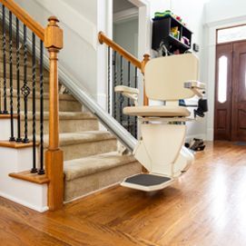 A beige stairlift is installed on a wooden staircase with carpeted steps and a dark metal railing in a residential hallway.