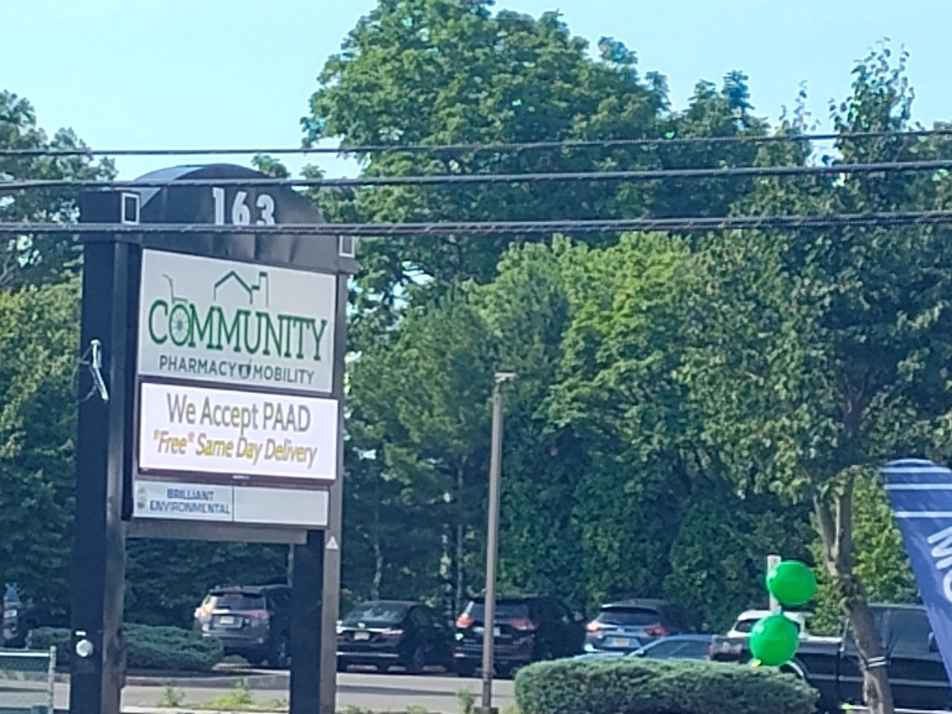 Sign for Community Pharmacy at 163, with trees and cars in the background. 