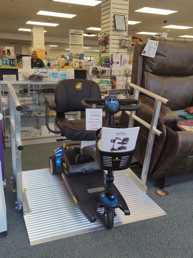 Mobility scooter on a ramp in a store; black, blue, and gold with a sign in front, brown recliner chair in background.