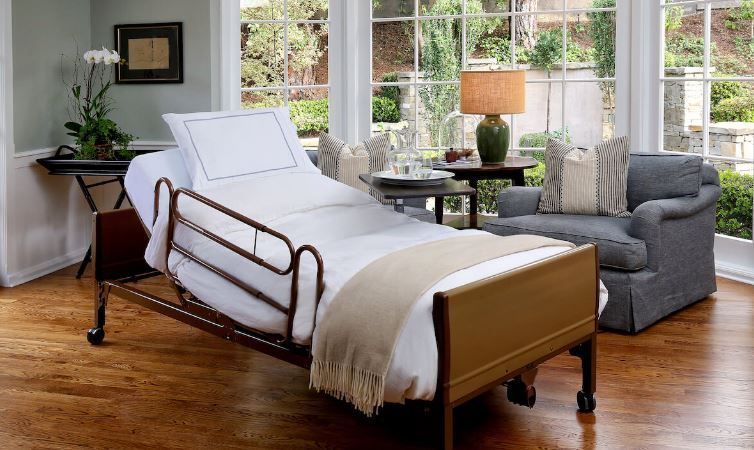 Hospital bed in a well-lit room with a cozy couch, side table, and large windows with outdoor view.