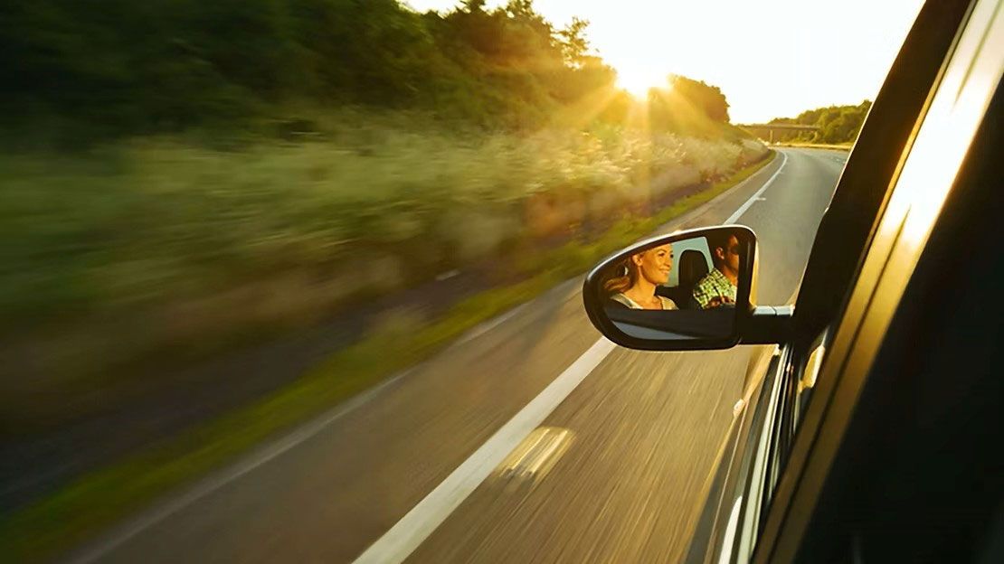 Car side mirror reflecting a person on a sunlit highway at dusk