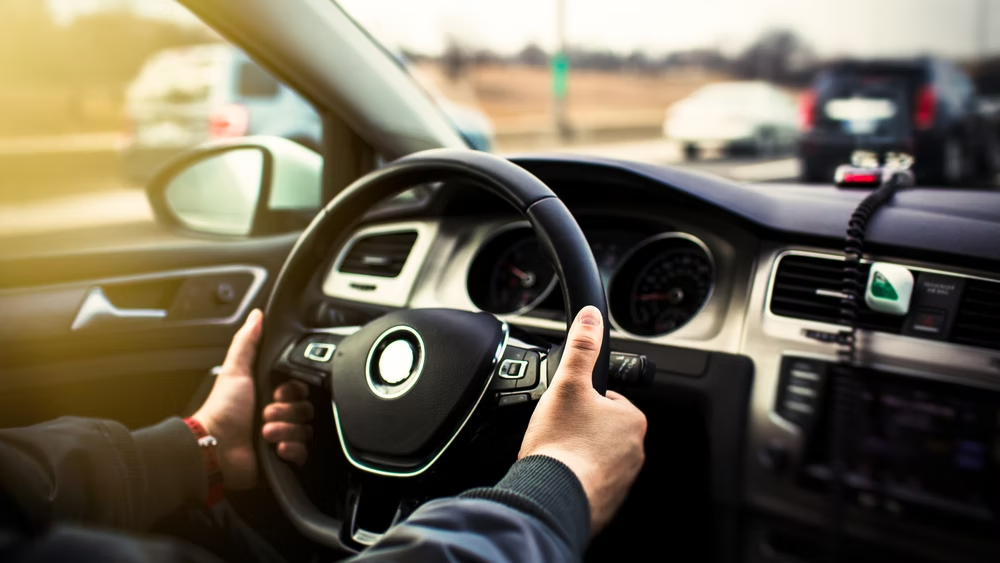 Driver holding the steering wheel in a car, with sunlight and traffic ahead.