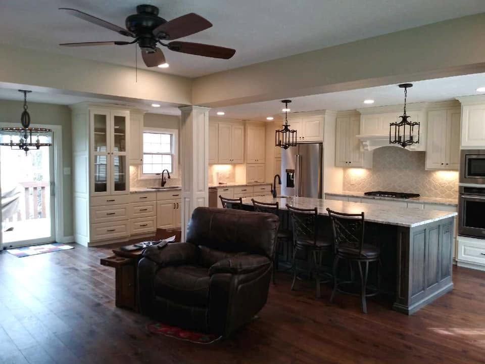 Open kitchen and living area with white cabinets, dark island stools, and a brown recliner under ceiling fans.