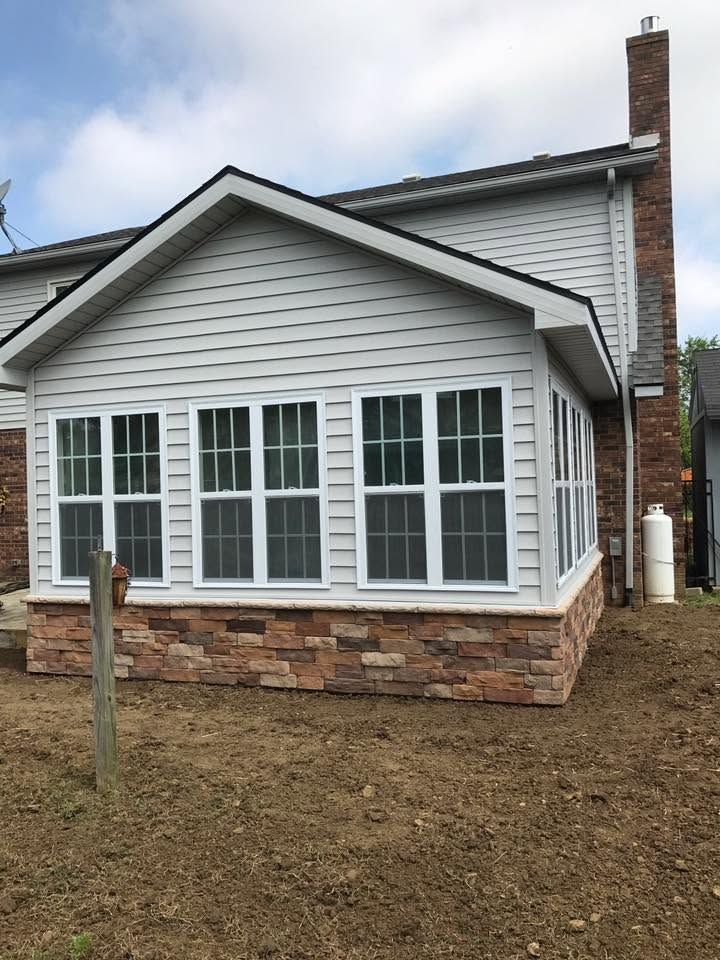 Single-story house exterior with white siding, stone base, and large front windows under a cloudy sky.