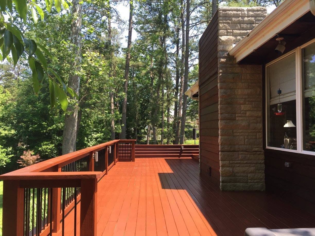 Wooden deck with red railing beside a brick house, overlooking a wooded backyard.