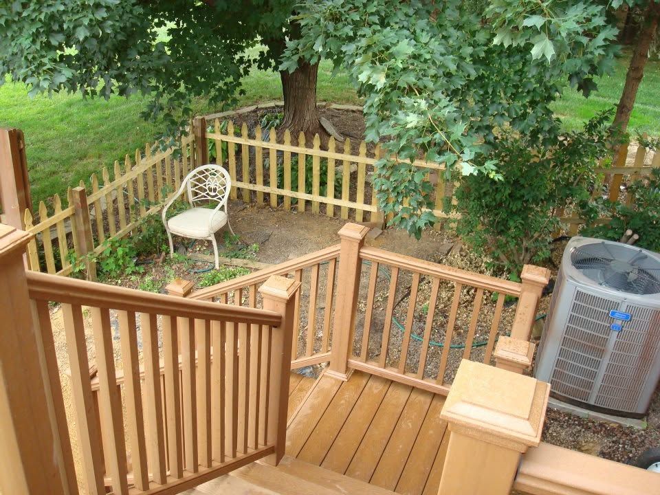 Wooden backyard deck and gate beside a fenced garden with a chair and air conditioner unit.