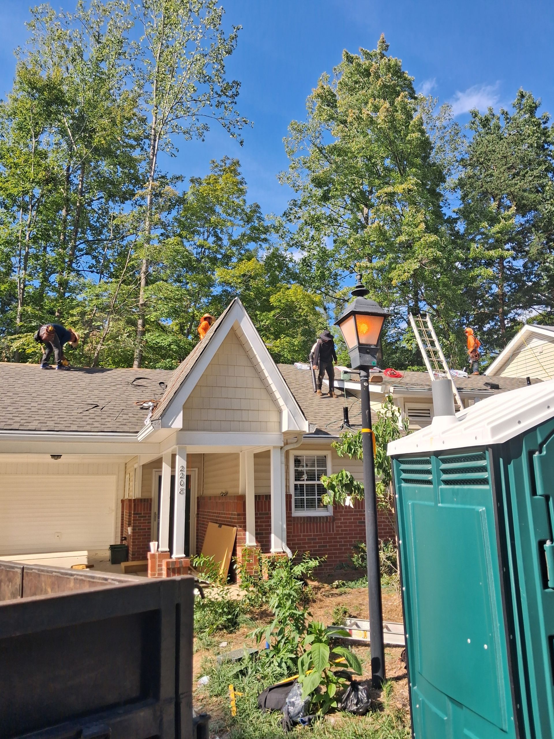 Two workers on a roof near a house with tall trees and a blue sky