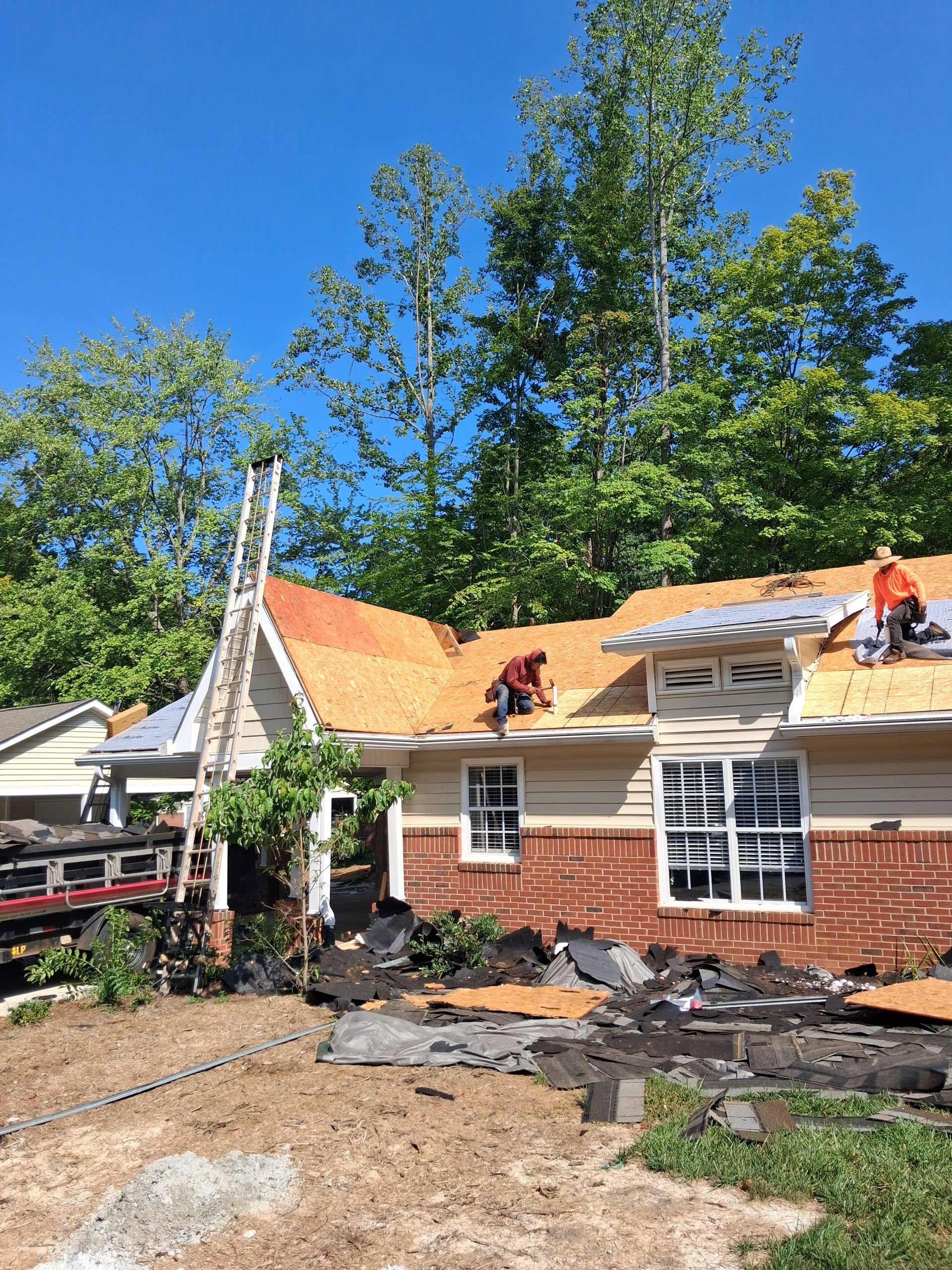 Workers repairing a damaged house roof with orange tarps under a clear blue sky